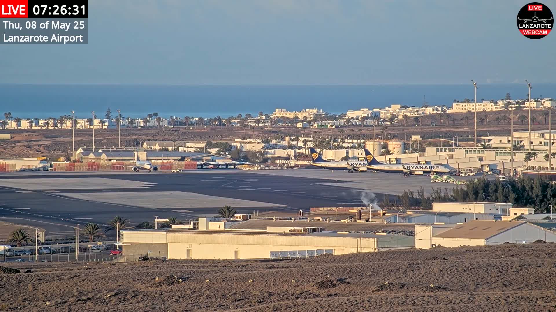 A clear, sunny day overlooks an airport tarmac with several airplanes, including two Ryanair planes, near buildings and the ocean.
