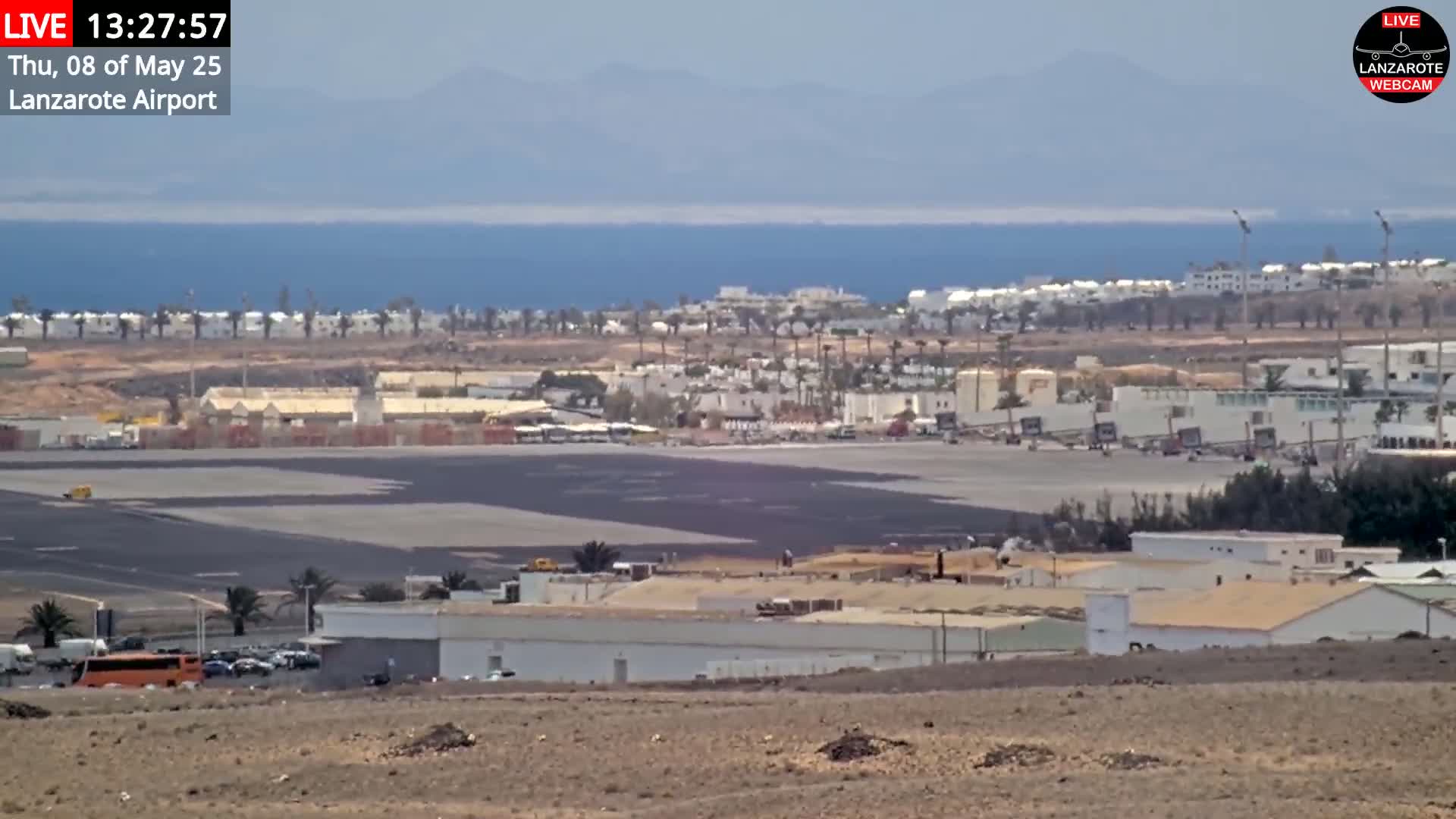 A long shot of an airport tarmac and surrounding buildings on a sunny day, with a calm ocean and distant mountains visible in the background.