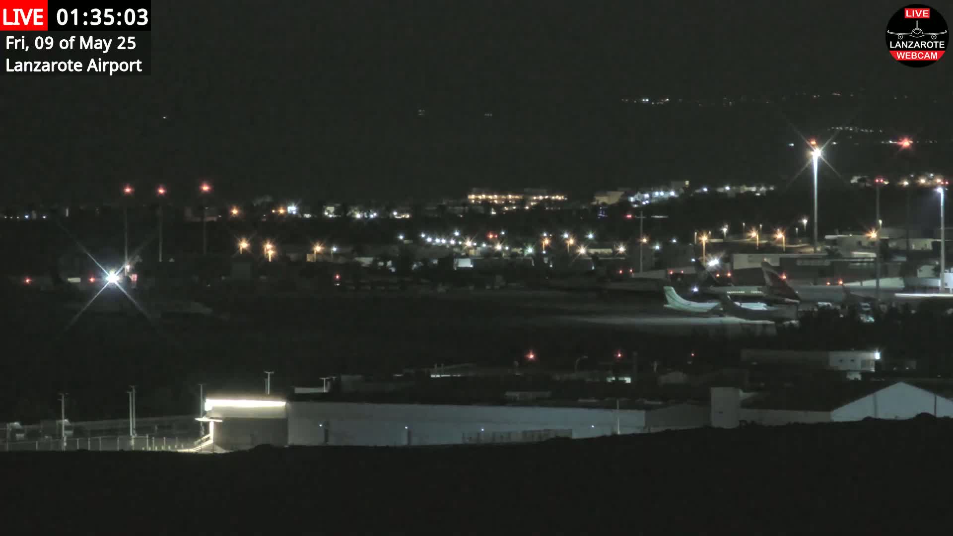 A nighttime long shot of an airport with several airplanes parked on the tarmac, surrounded by buildings and lights under a clear, dark sky.