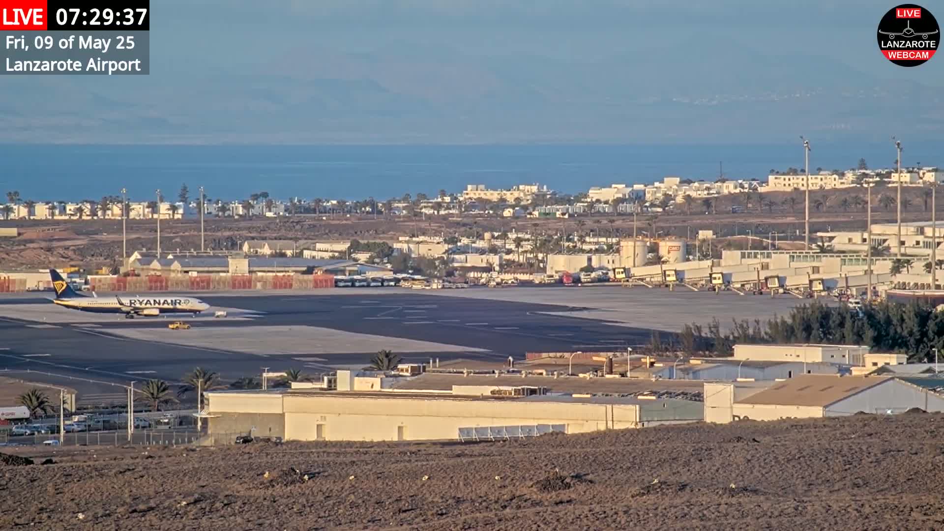 A Ryanair airplane sits on a tarmac at an airport overlooking a town and calm ocean under a clear sky.
