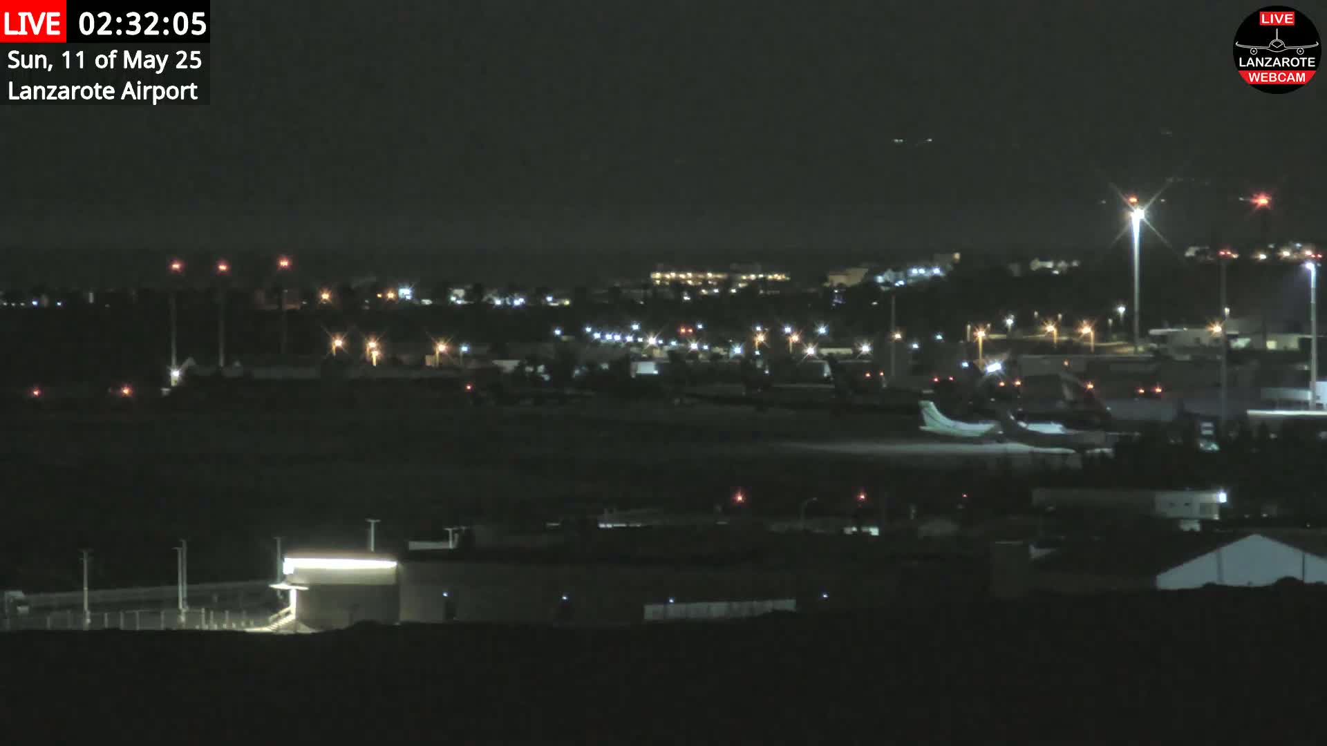 A nighttime long shot of an airport runway with a plane on the tarmac and various lights illuminating the surrounding buildings and landscape.