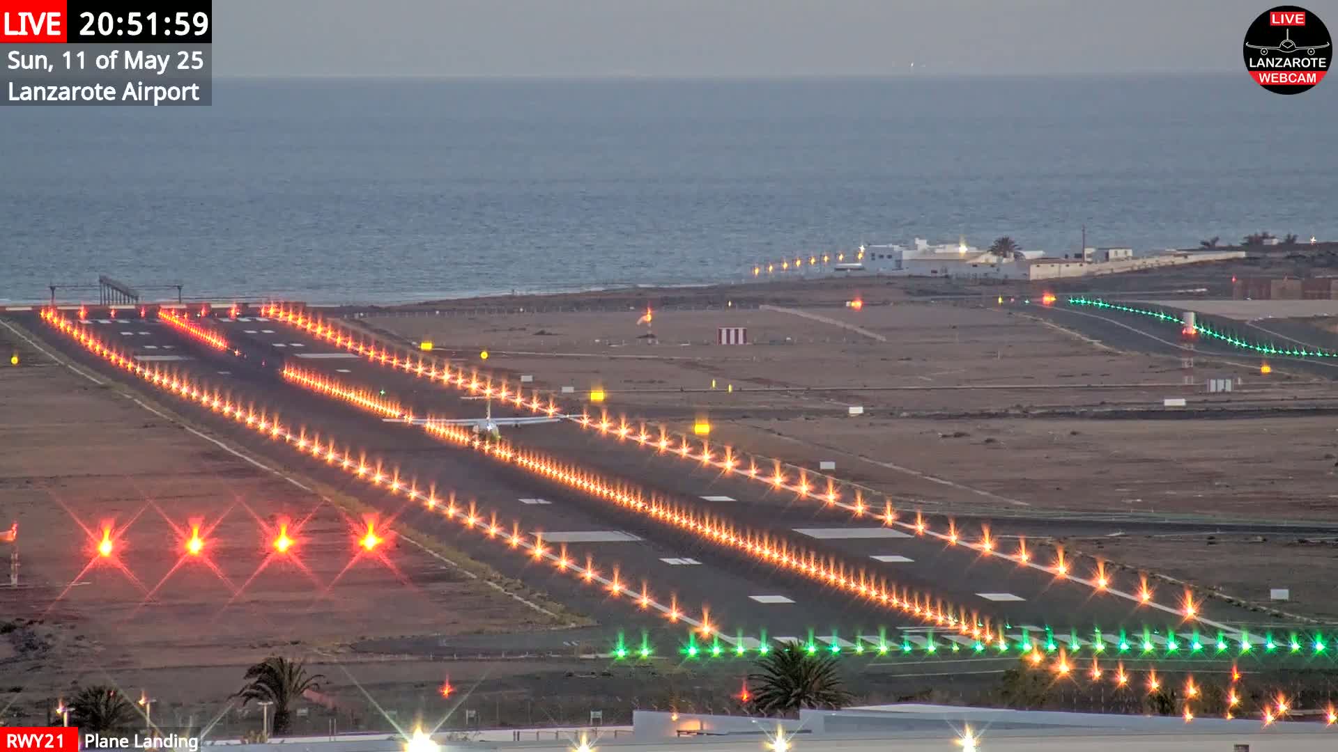 A plane is landing on a brightly lit runway at an airport next to a calm ocean at dusk.
