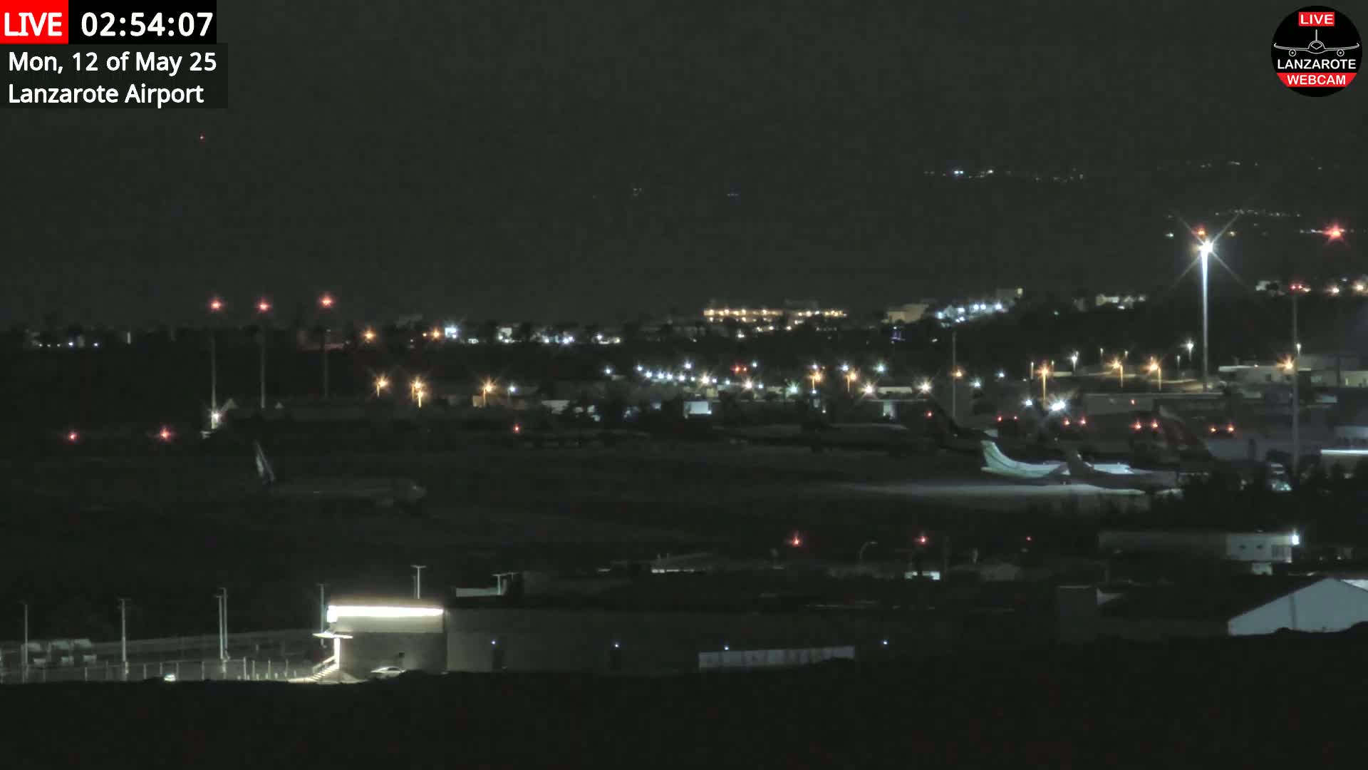 A nighttime view of an airport tarmac with several planes, buildings, and lights, under a clear, dark sky.