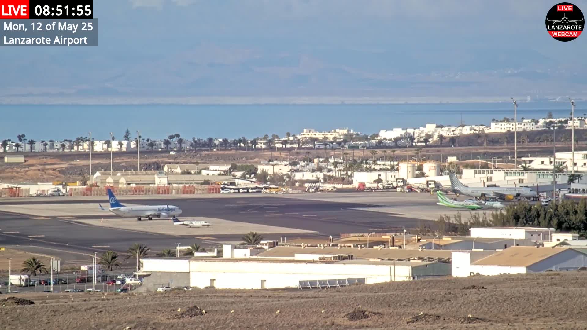 A sunny day shows an airport tarmac with several airplanes, airport buildings, and a body of water in the background.