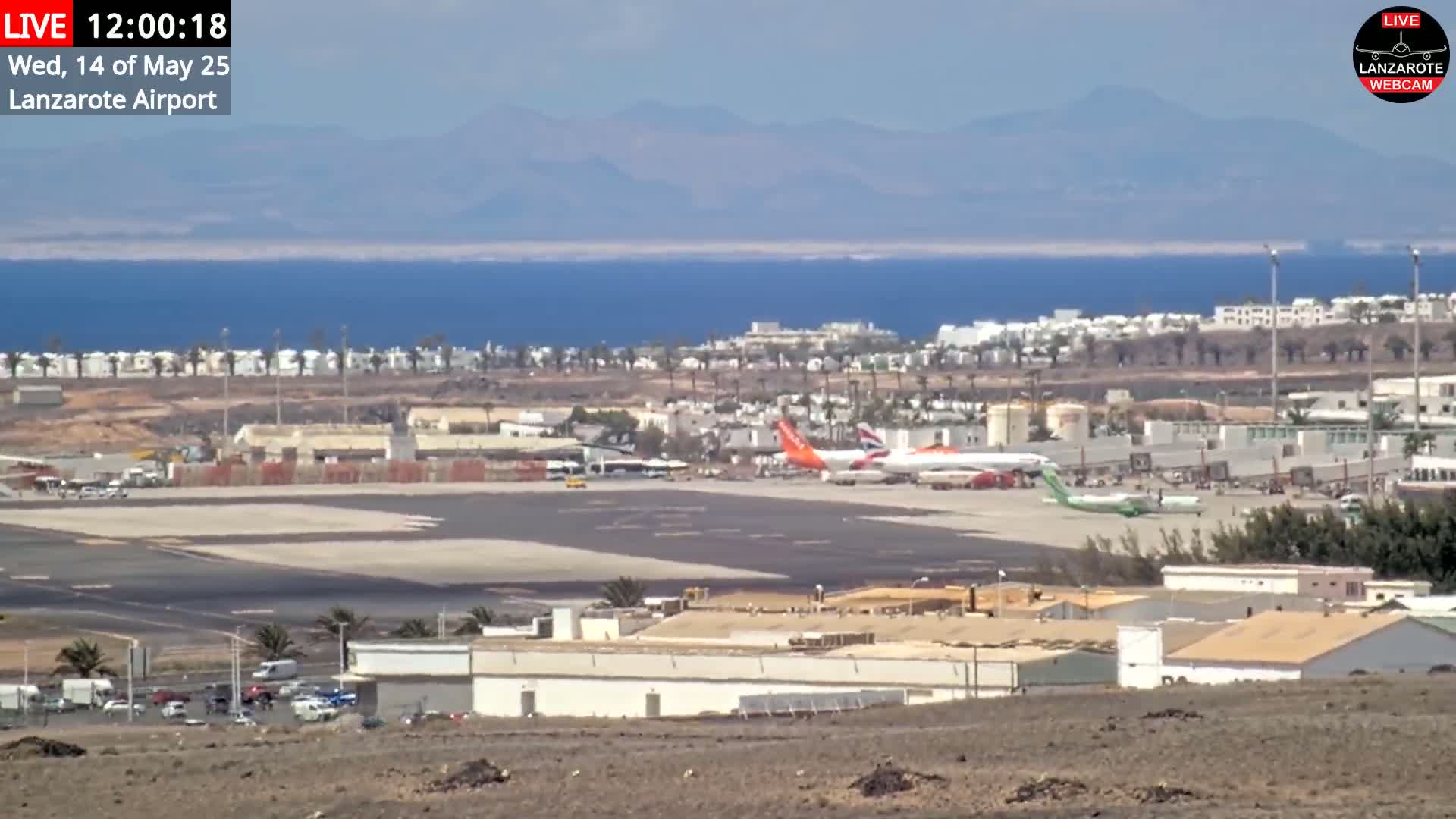 A sunny day at an airport shows several airplanes parked on the tarmac near buildings and the ocean, with mountains visible in the distance.