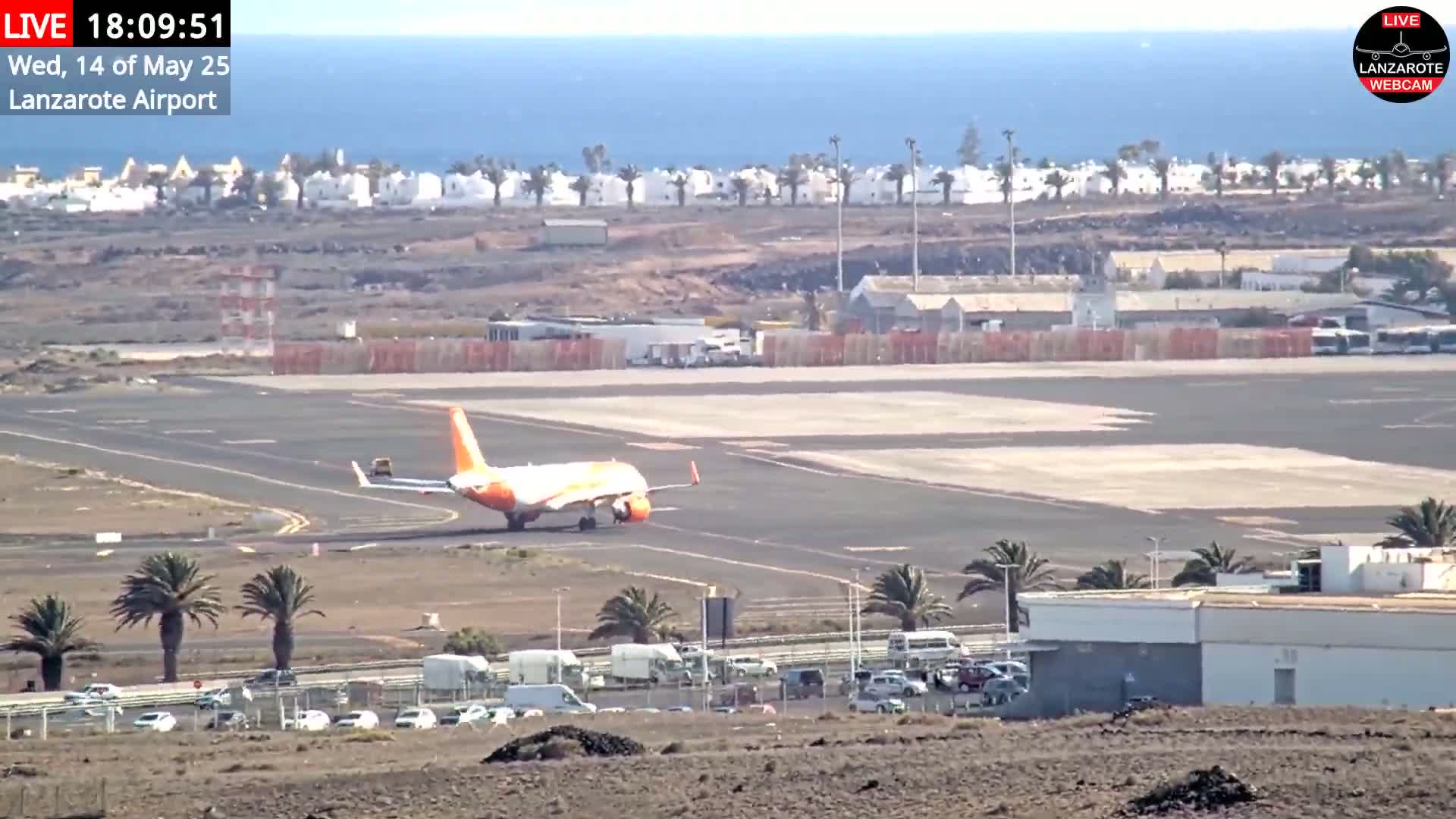 An orange-tailed airplane taxis on an airport runway near a coastline under a clear sky.