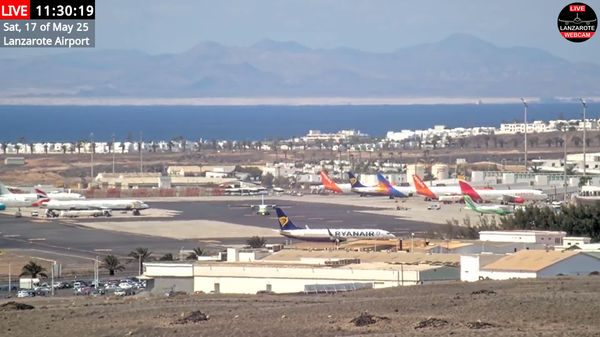 Several airplanes are parked on a tarmac at an airport by the ocean under a sunny sky, with mountains visible in the distance.