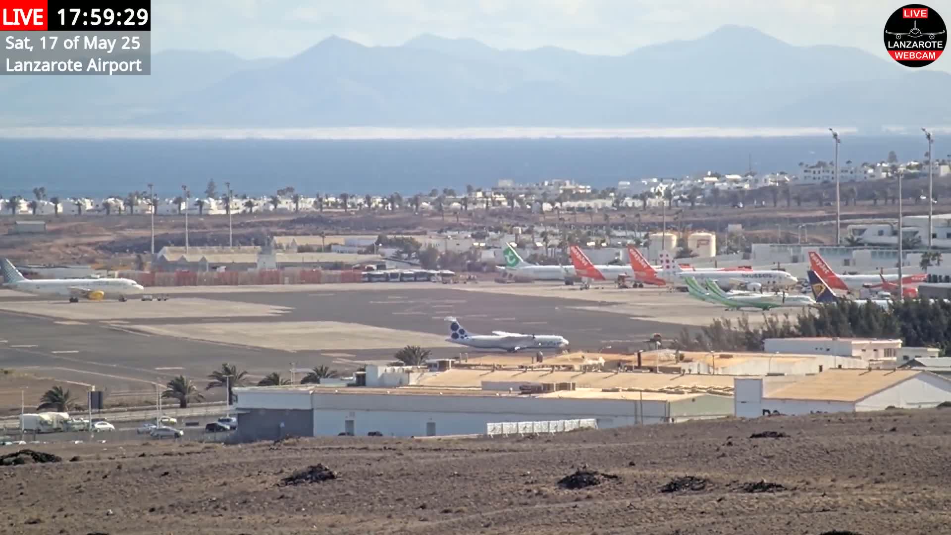 A sunny day at an airport shows several airplanes parked on the tarmac, with a view of the ocean and distant mountains.