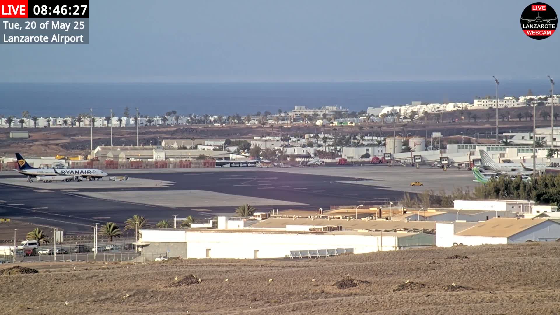 An airport tarmac with several airplanes, airport buildings, and a view of the ocean under a clear sky.