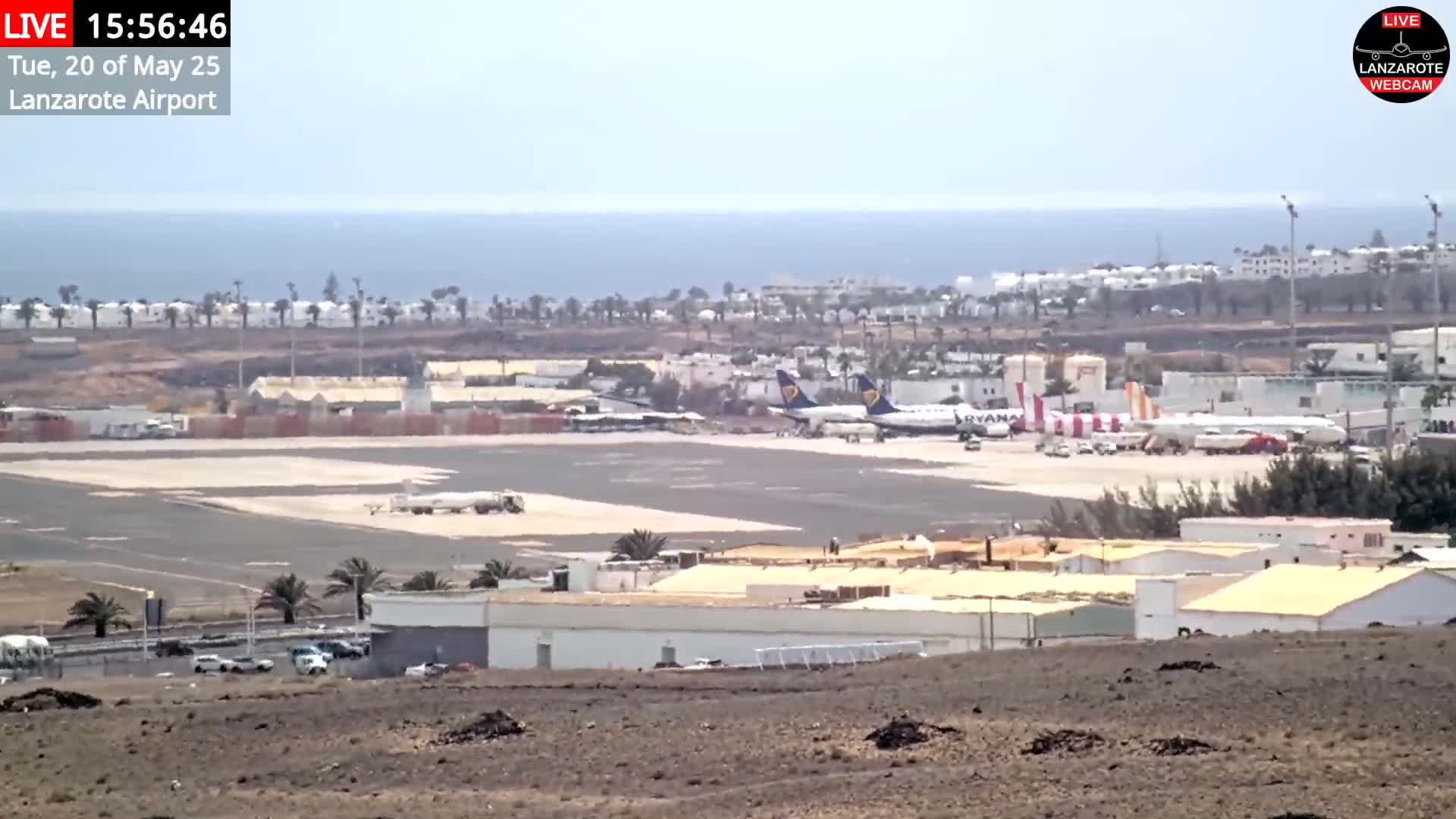 A sunny day at an airport shows several airplanes on the tarmac, surrounded by buildings and palm trees, with the ocean visible in the background.