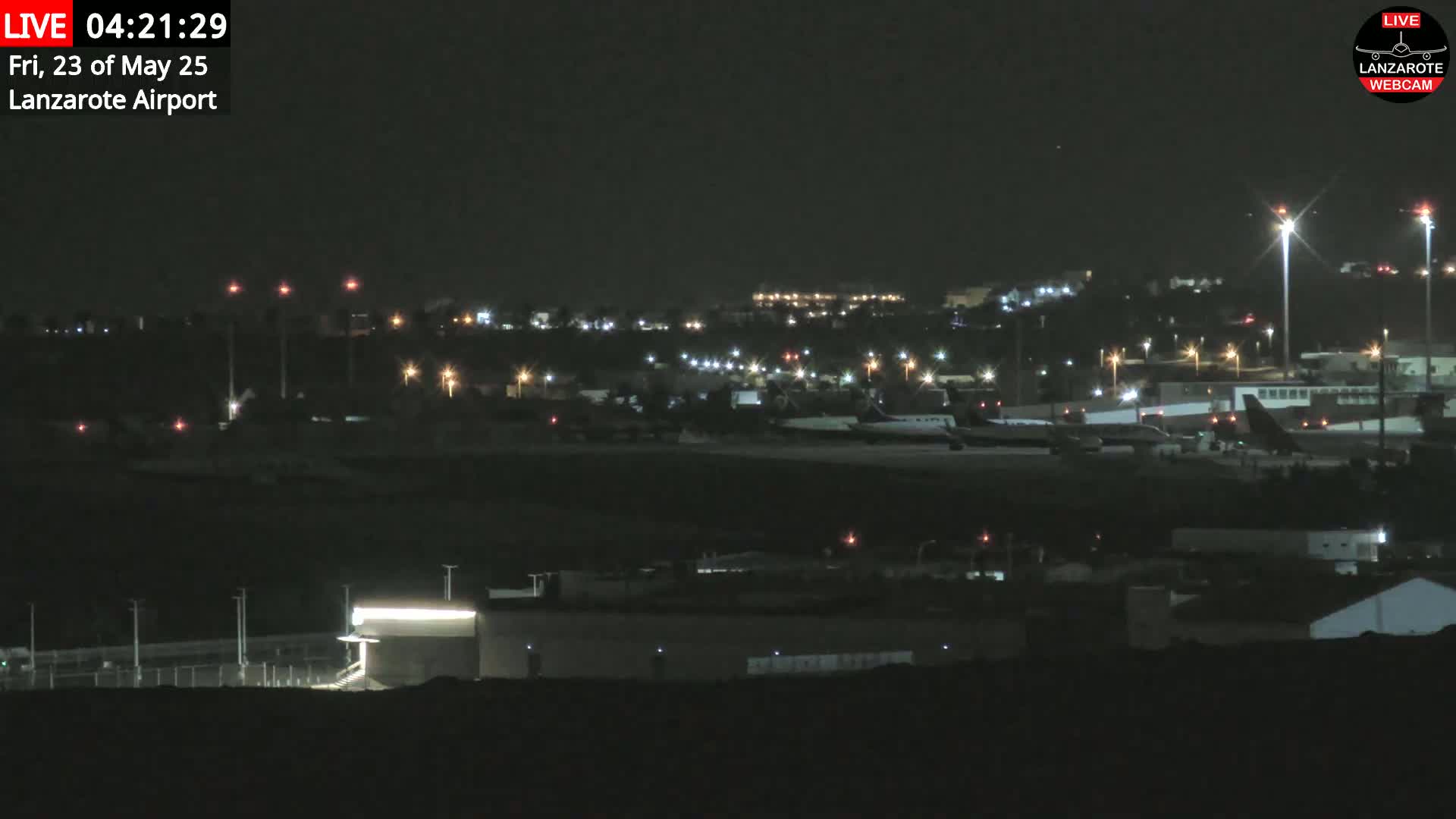 A nighttime long shot of an airport tarmac with several airplanes parked, illuminated by numerous lights under a dark sky.
