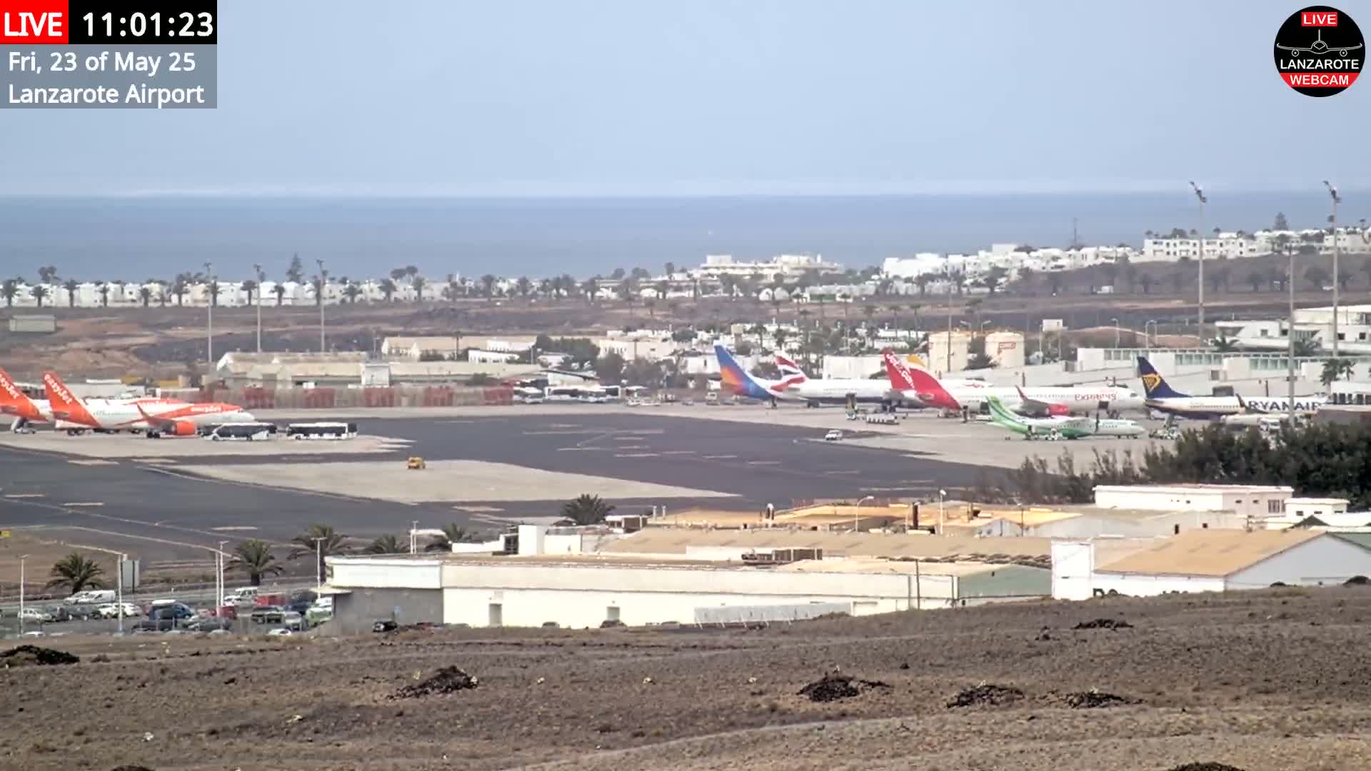 A distant view of an airport with several airplanes parked on the tarmac, under a hazy sky, with the ocean visible in the background.