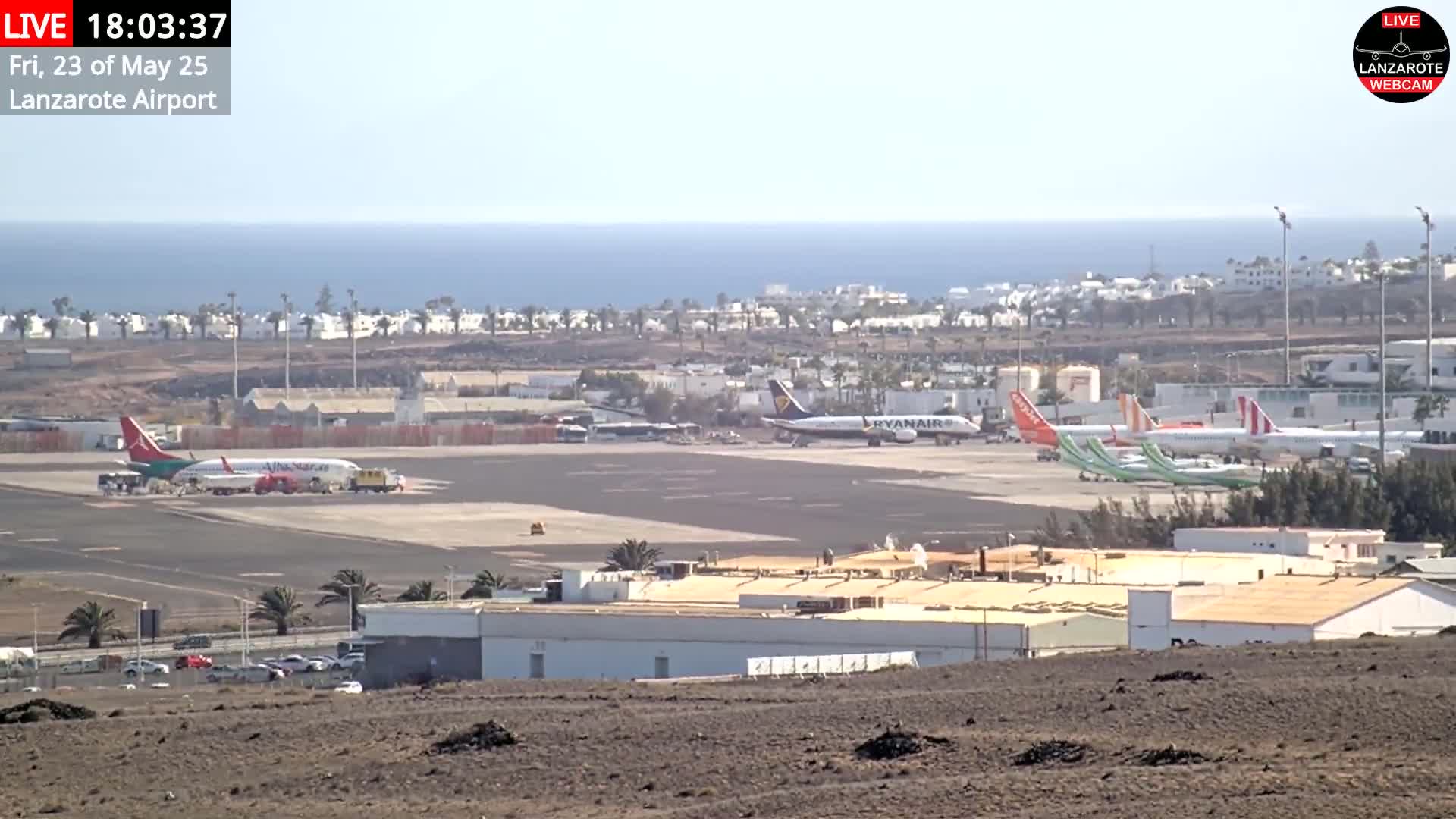 A sunny day at an airport shows several airplanes parked on the tarmac near buildings, with the ocean visible in the background.