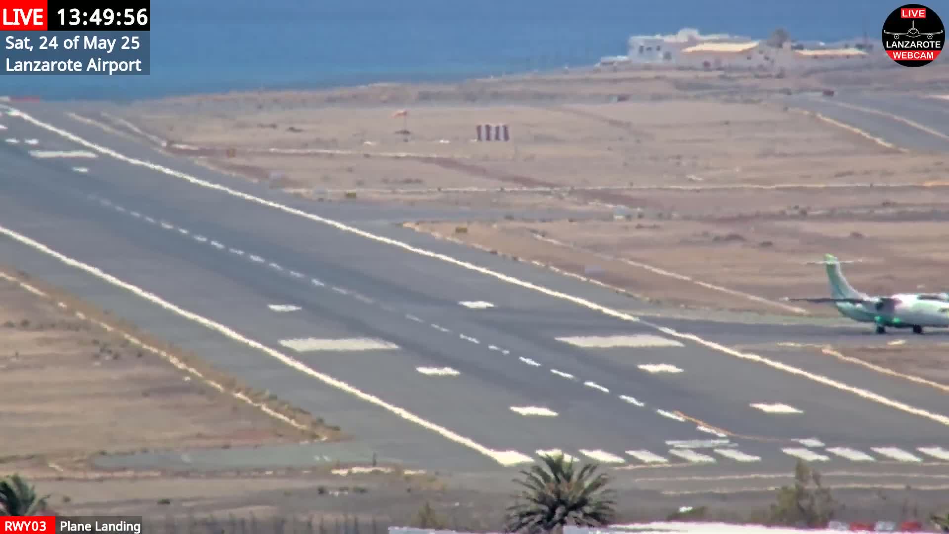A small airplane is landing on a runway at an airport, with a clear, sunny day and ocean visible in the background.
