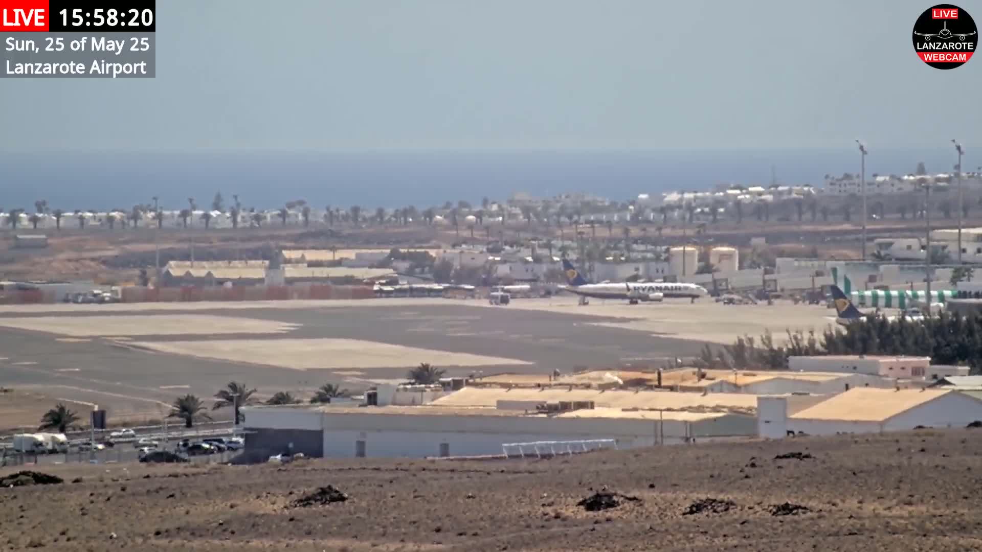 A sunny day at an airport shows several airplanes on the tarmac, buildings, and palm trees, with the ocean visible in the distance.