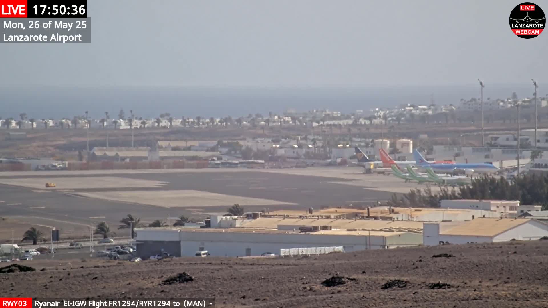 A hazy view of an airport tarmac with several airplanes parked, buildings, and a distant ocean under a light haze.
