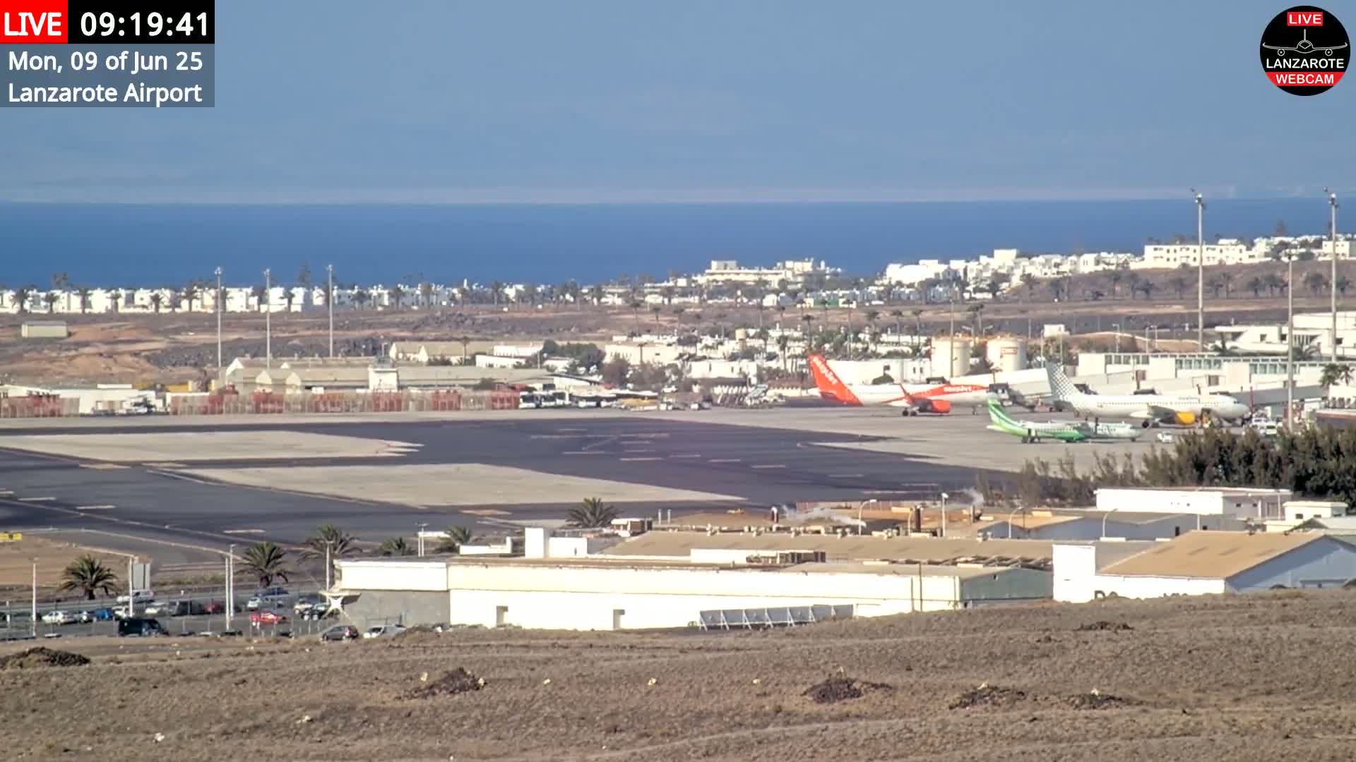 A sunny day at an airport shows several airplanes parked on the tarmac near buildings, with a calm ocean in the background.