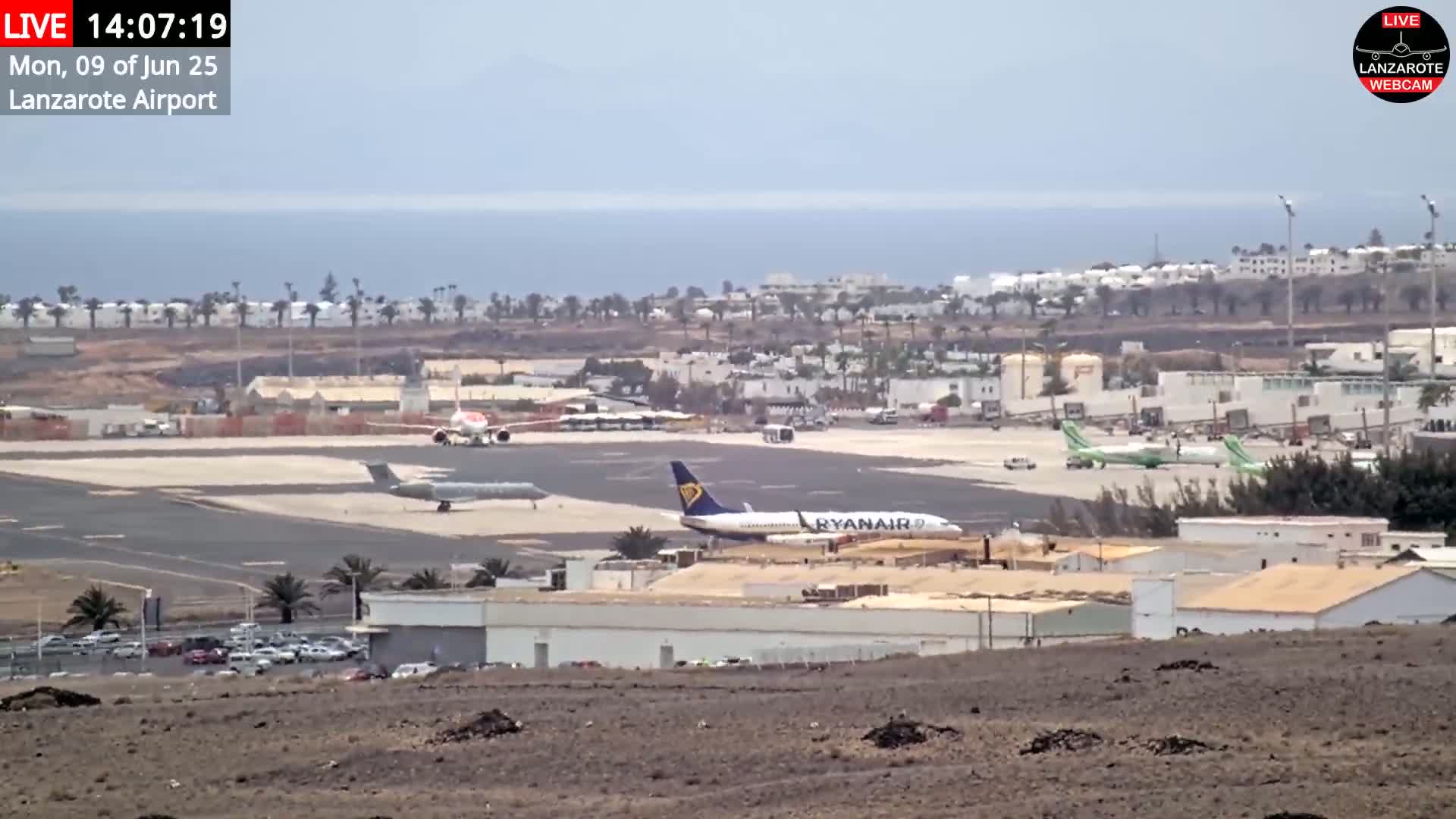 A high-angle view of an airport tarmac with several airplanes, including a Ryanair plane, under mostly clear skies with a calm ocean in the background.
