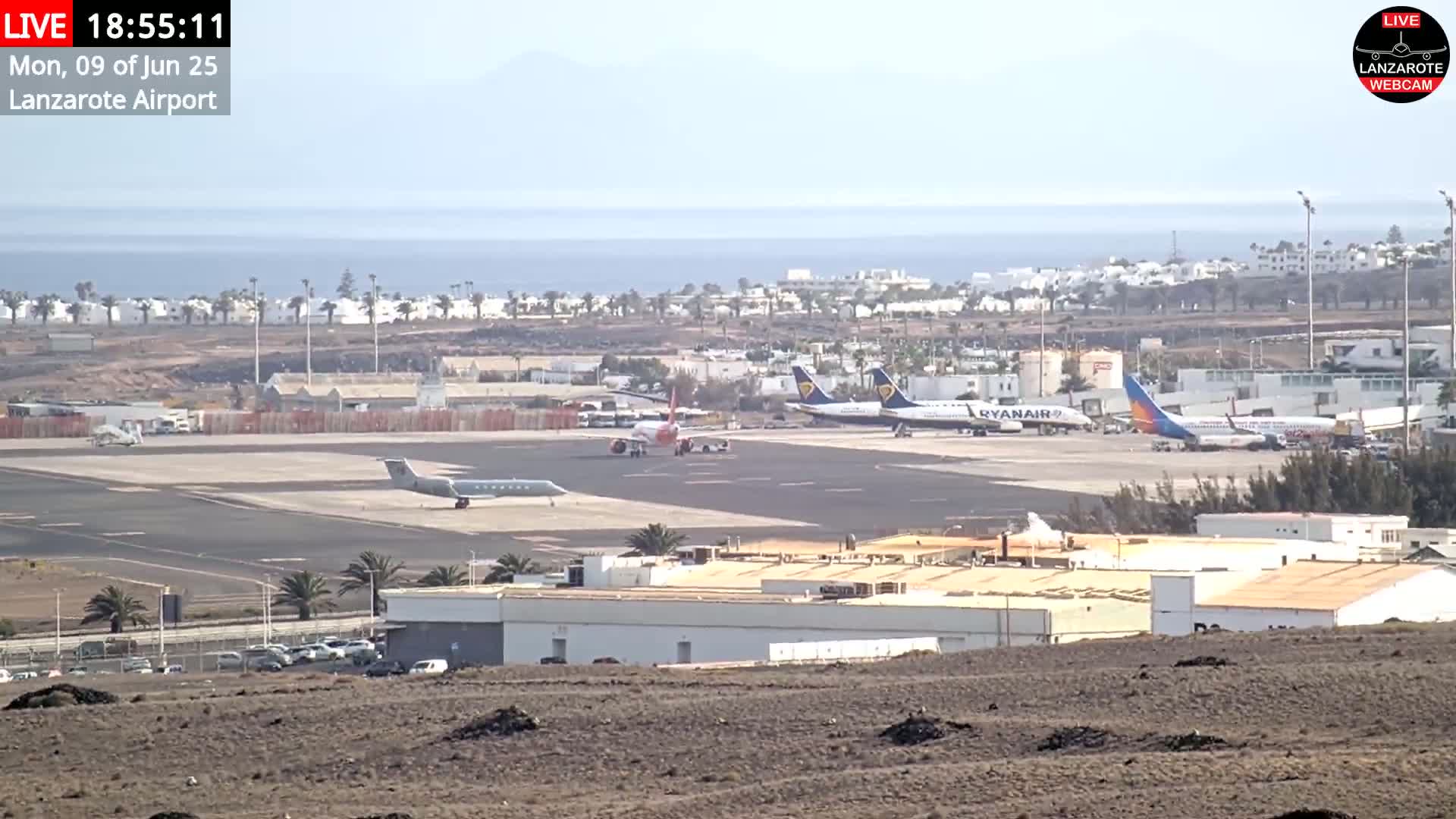 A sunny, distant view of an airport tarmac with several airplanes, airport buildings, and a coastline visible in the background.