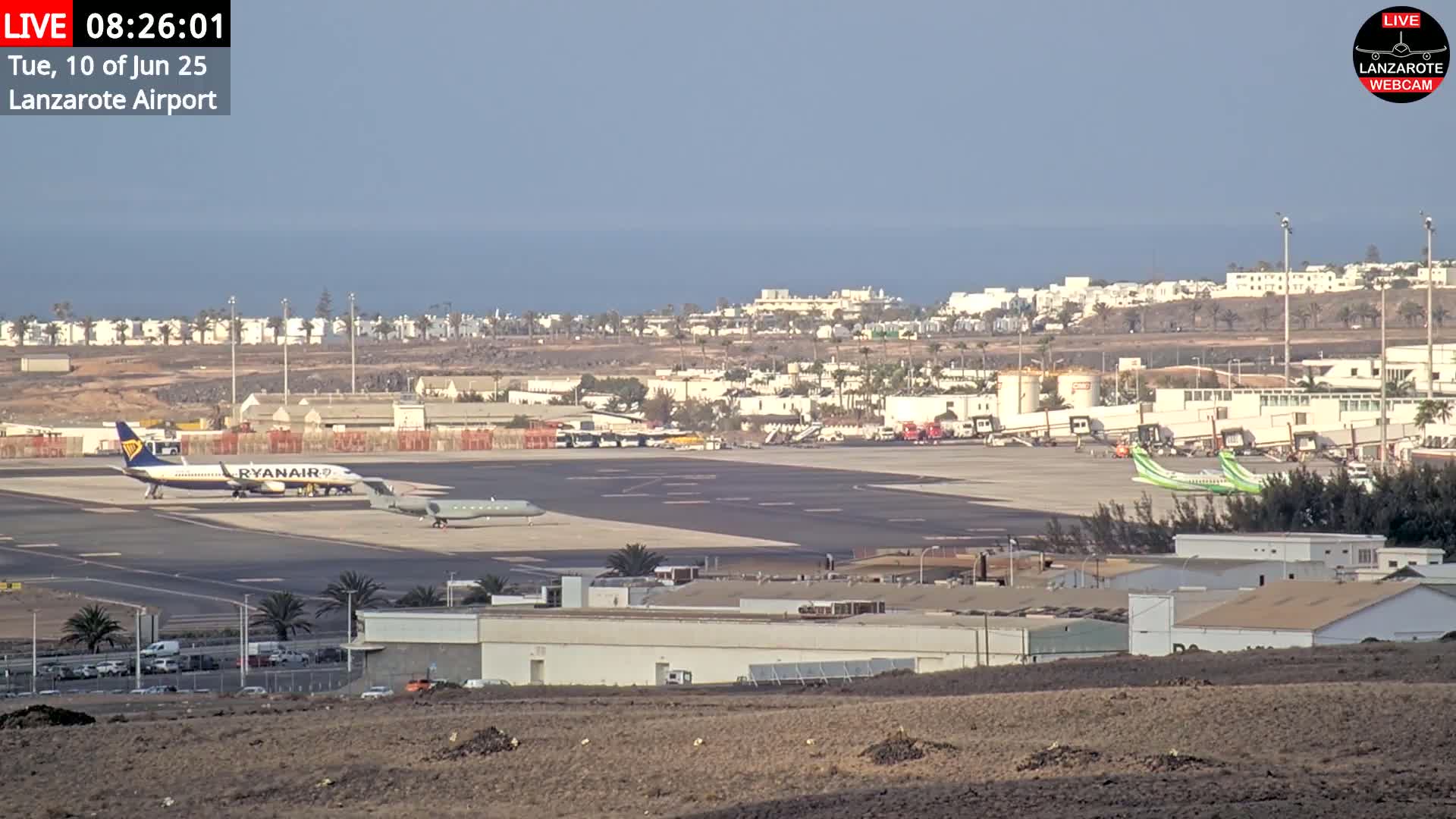 A sunny airport tarmac with several airplanes, airport buildings, and a distant ocean view.