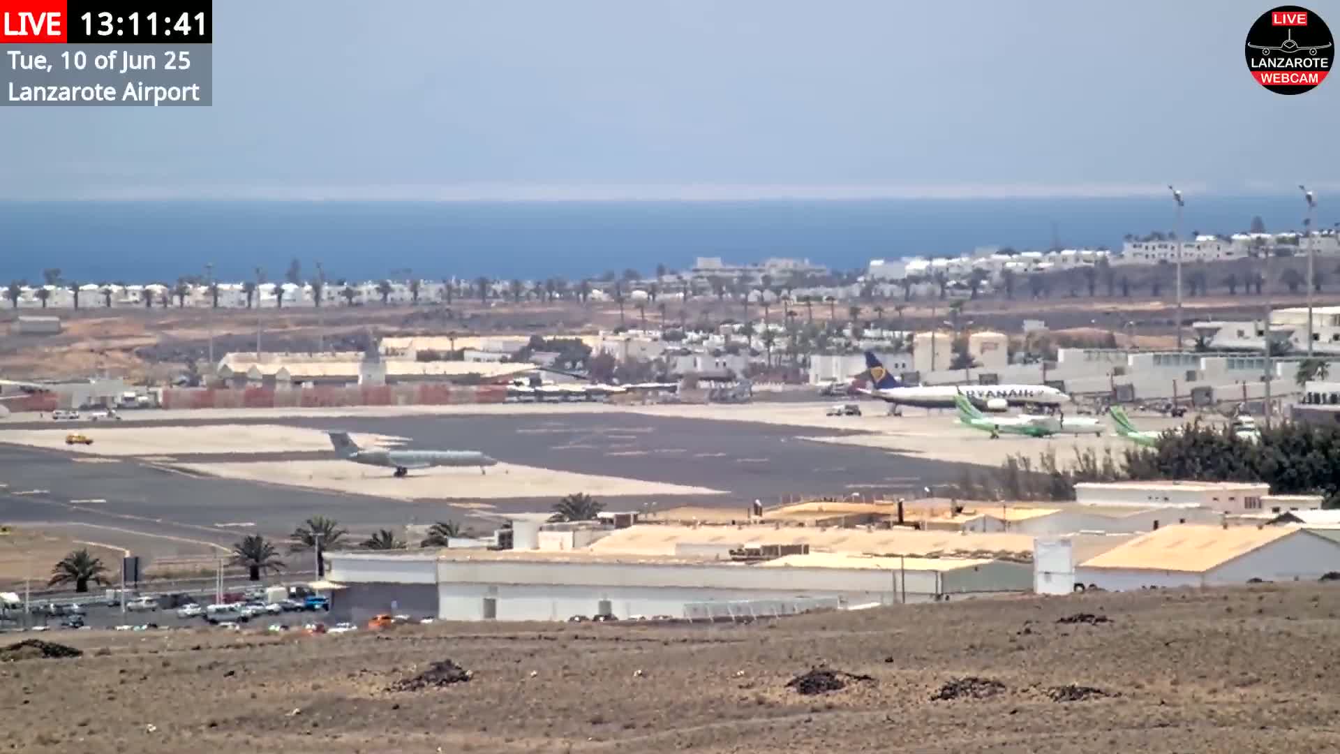 A long shot of an airport tarmac with several airplanes, surrounded by buildings and a coastline under sunny skies.