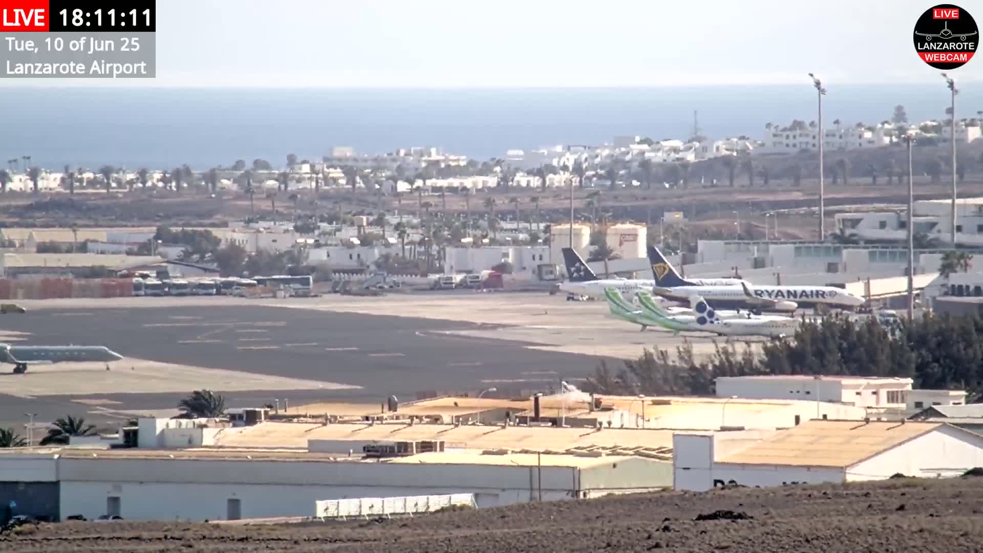 A sunny day at an airport shows several airplanes parked on the tarmac near buildings and a calm ocean in the background.