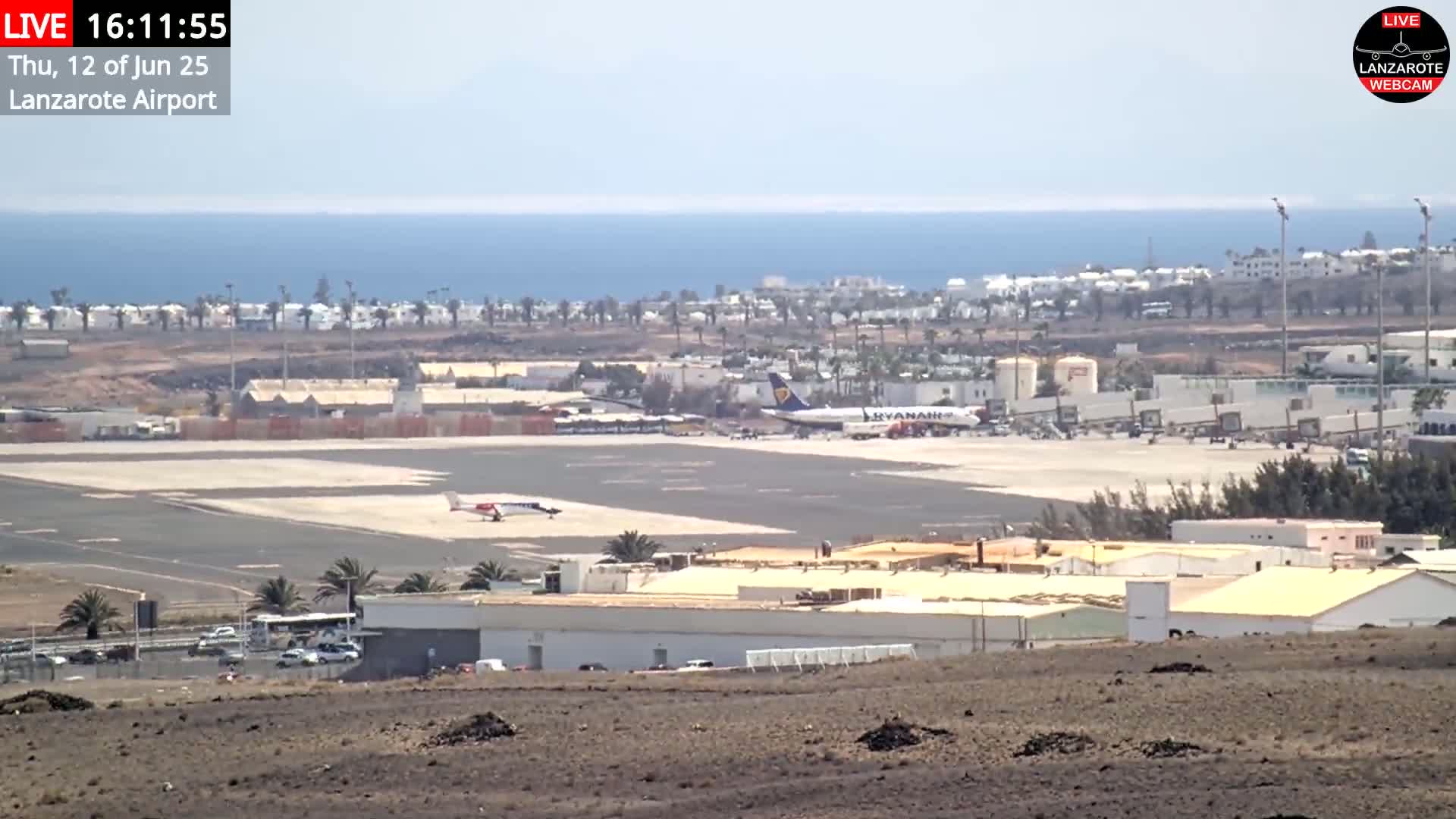 A sunny day at an airport shows a large passenger plane and a smaller plane on the tarmac, with the ocean visible in the background.