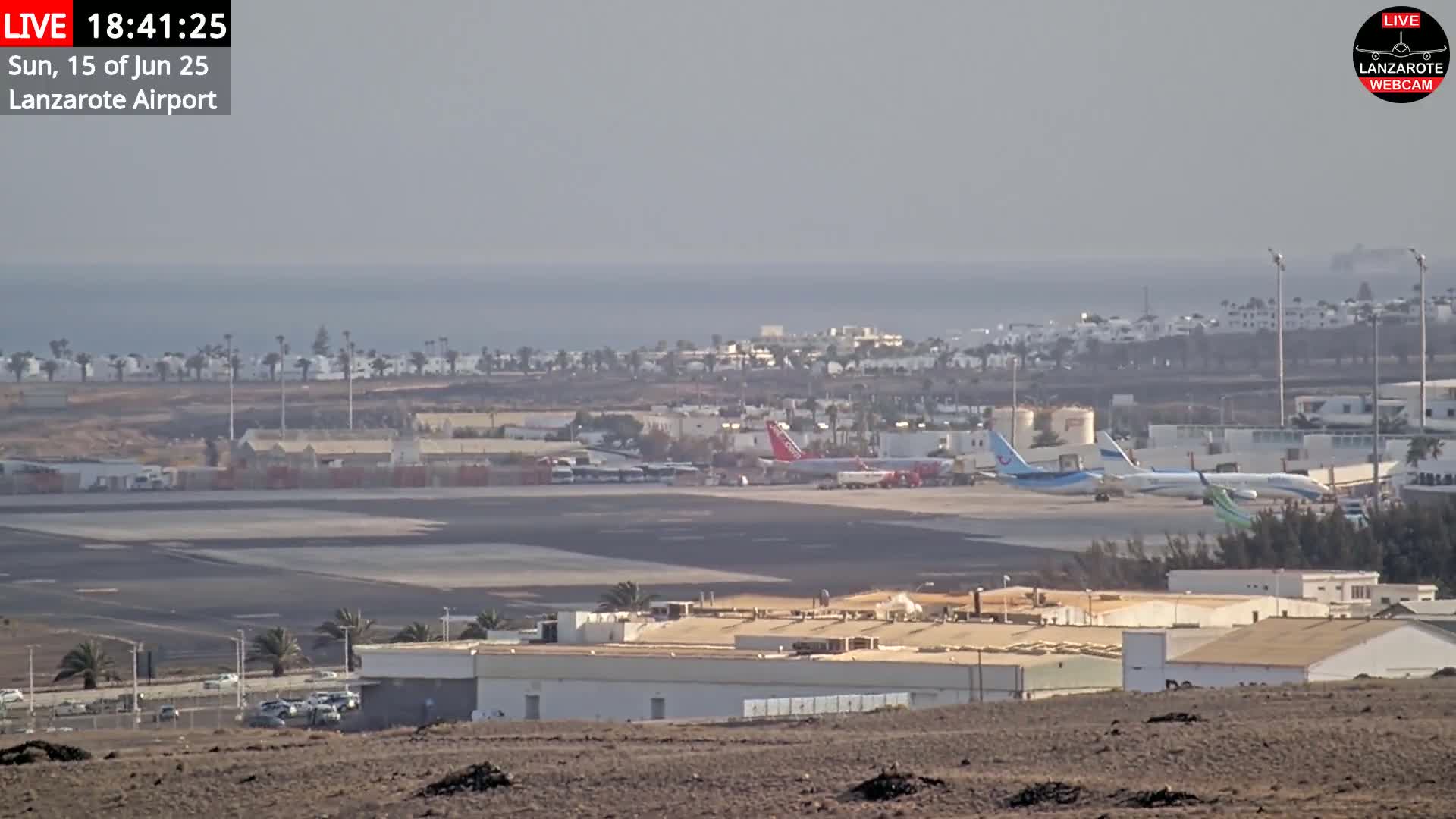 A hazy view of an airport with several airplanes on the tarmac, buildings, and a calm ocean in the distance.