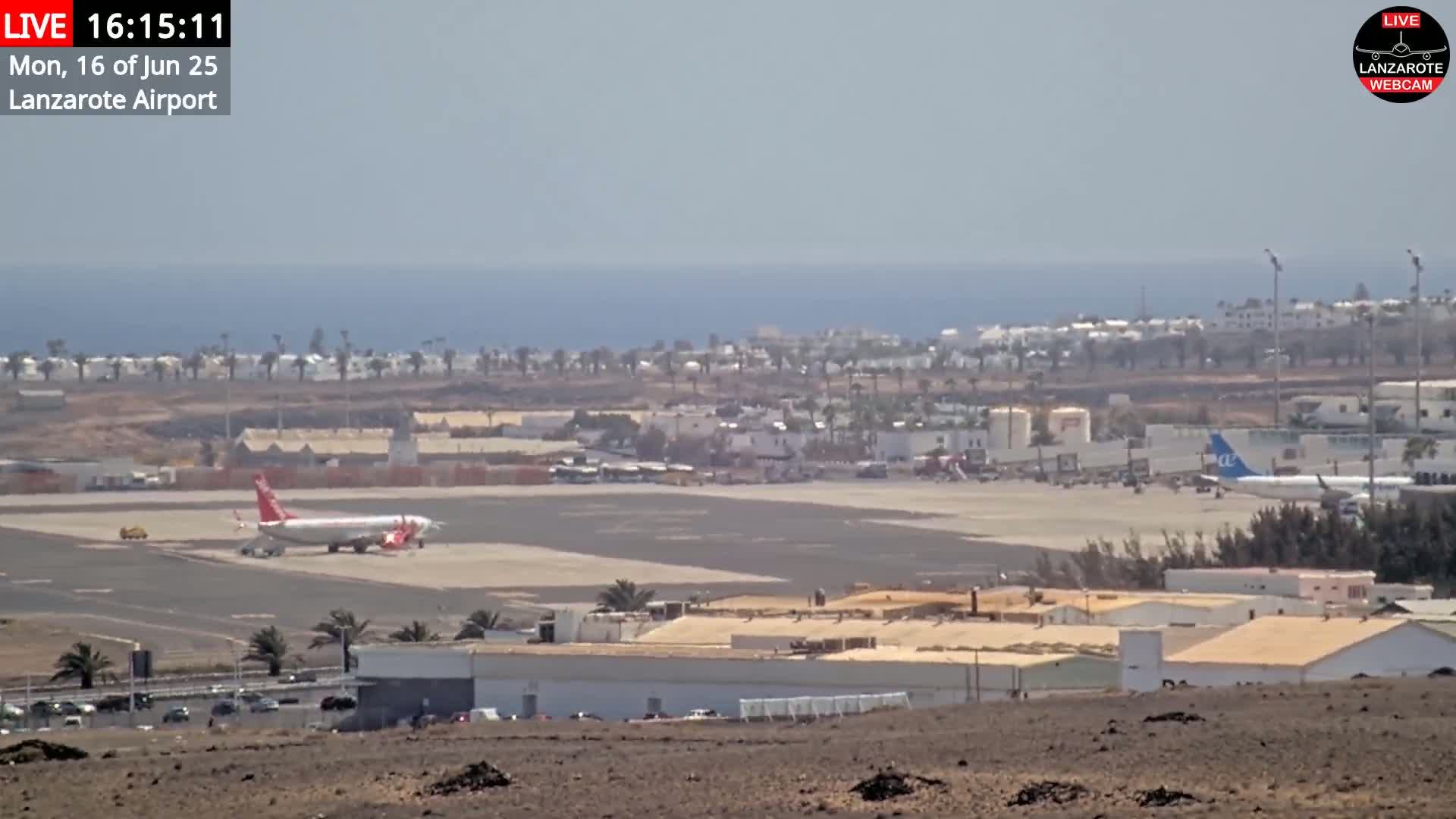 A long shot of an airport on a sunny day with a red and white airplane on the tarmac, buildings and a coastline in the background.