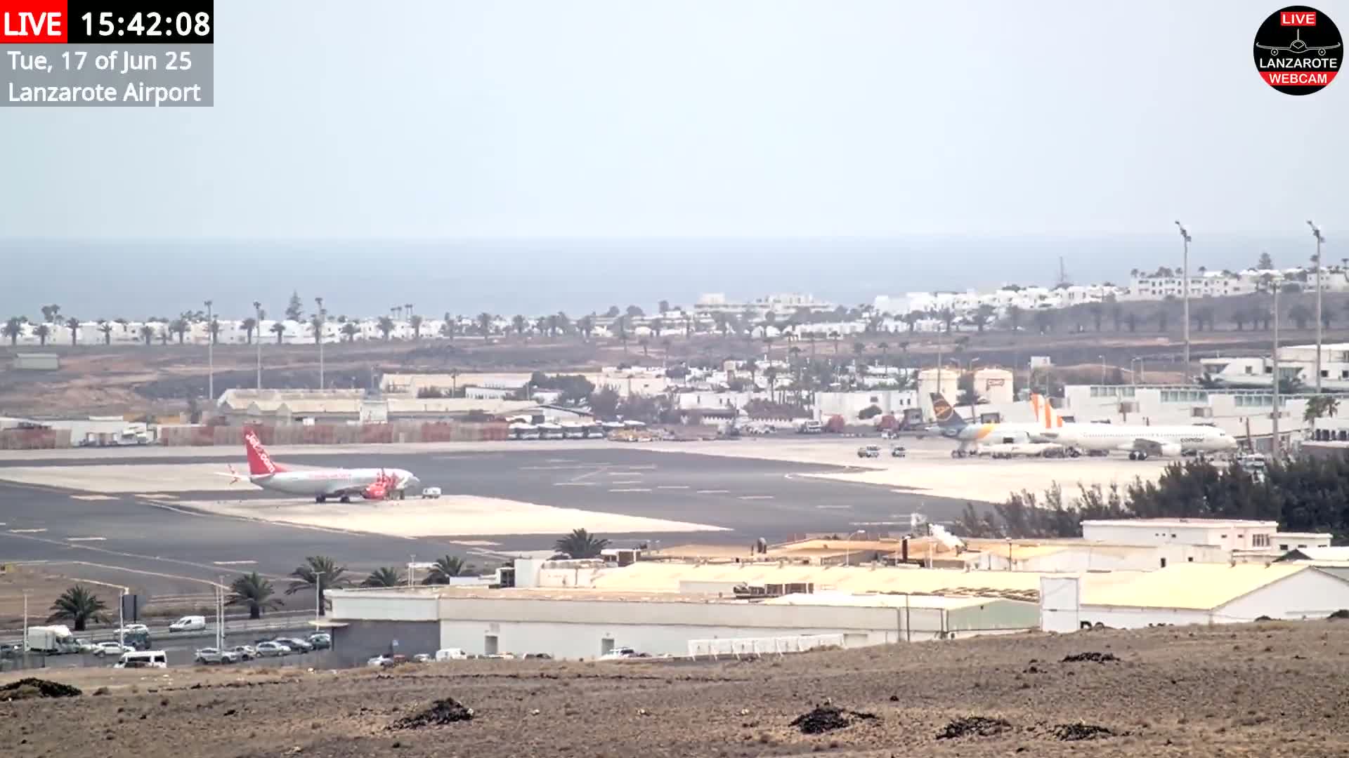 An airport tarmac with several planes, including one parked and another on the runway, is visible under hazy skies, with an ocean and buildings in the background.
