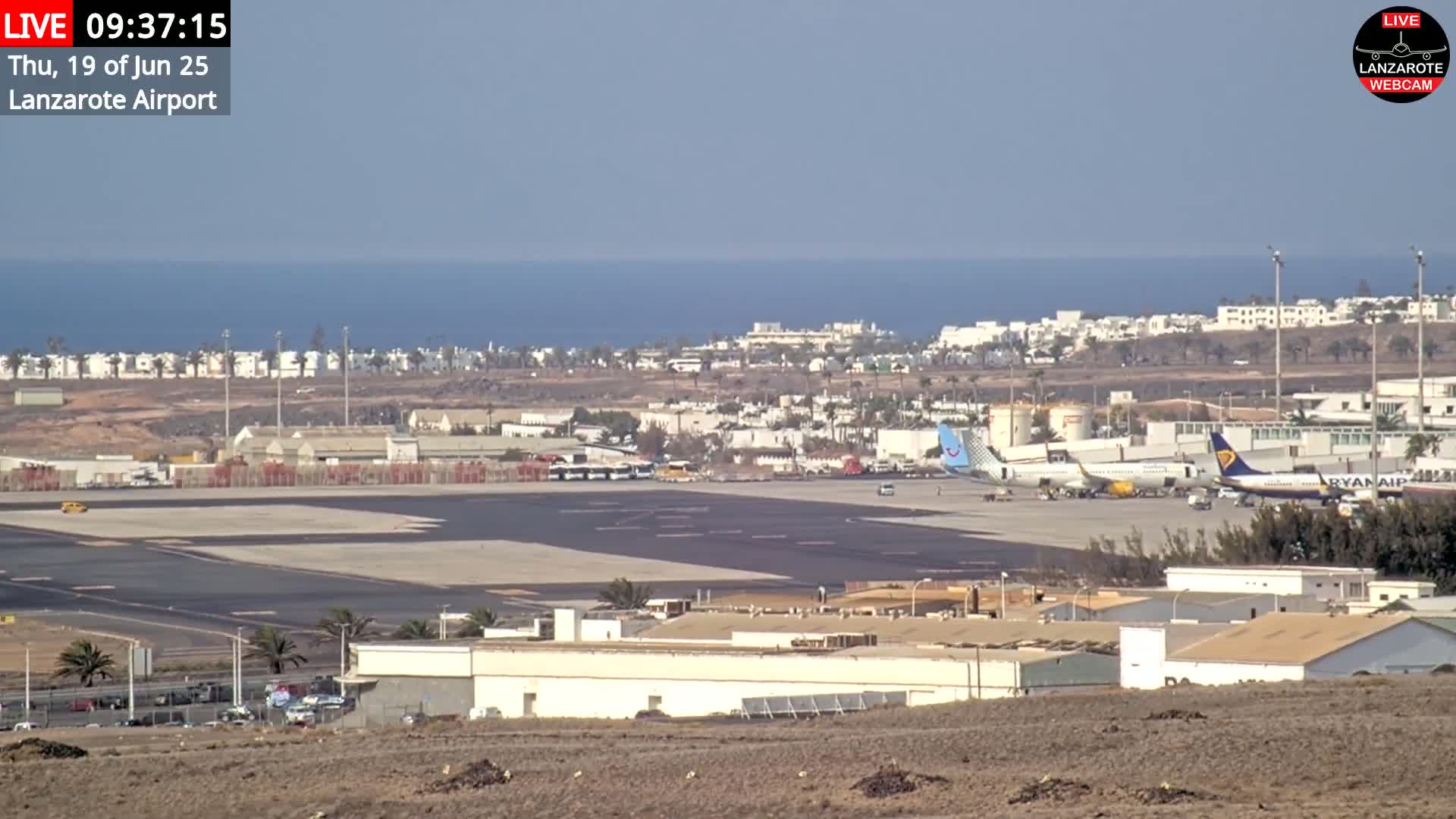 An airport tarmac with several airplanes parked, adjacent buildings, and a distant ocean under a clear sky.