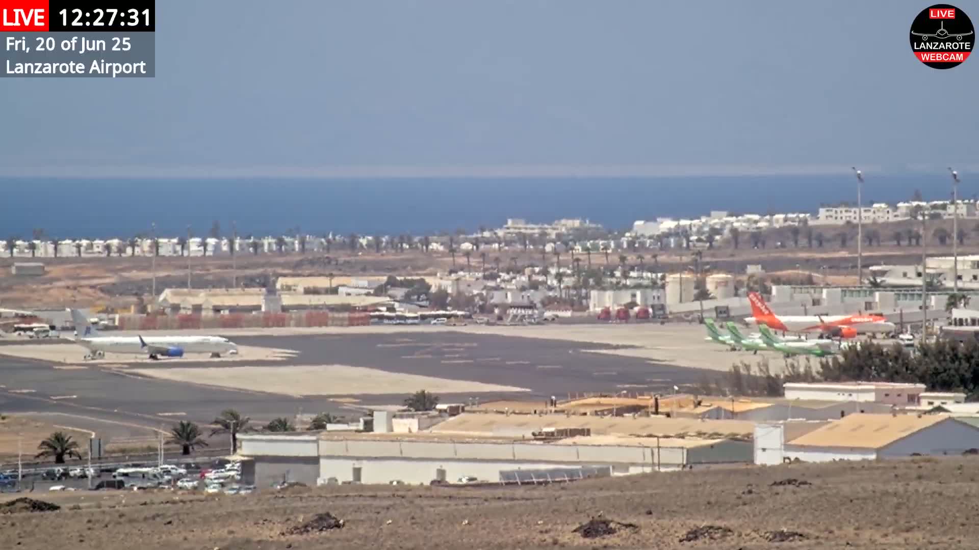 A long shot of an airport tarmac with several airplanes and surrounding buildings on a sunny day, overlooking a calm ocean in the distance.