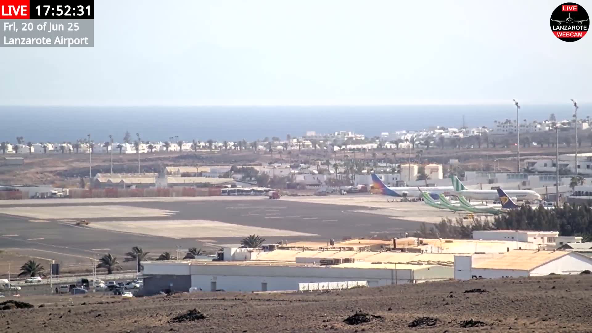 A long shot of an airport tarmac with several airplanes parked, adjacent buildings, and a calm ocean in the background on a clear day.