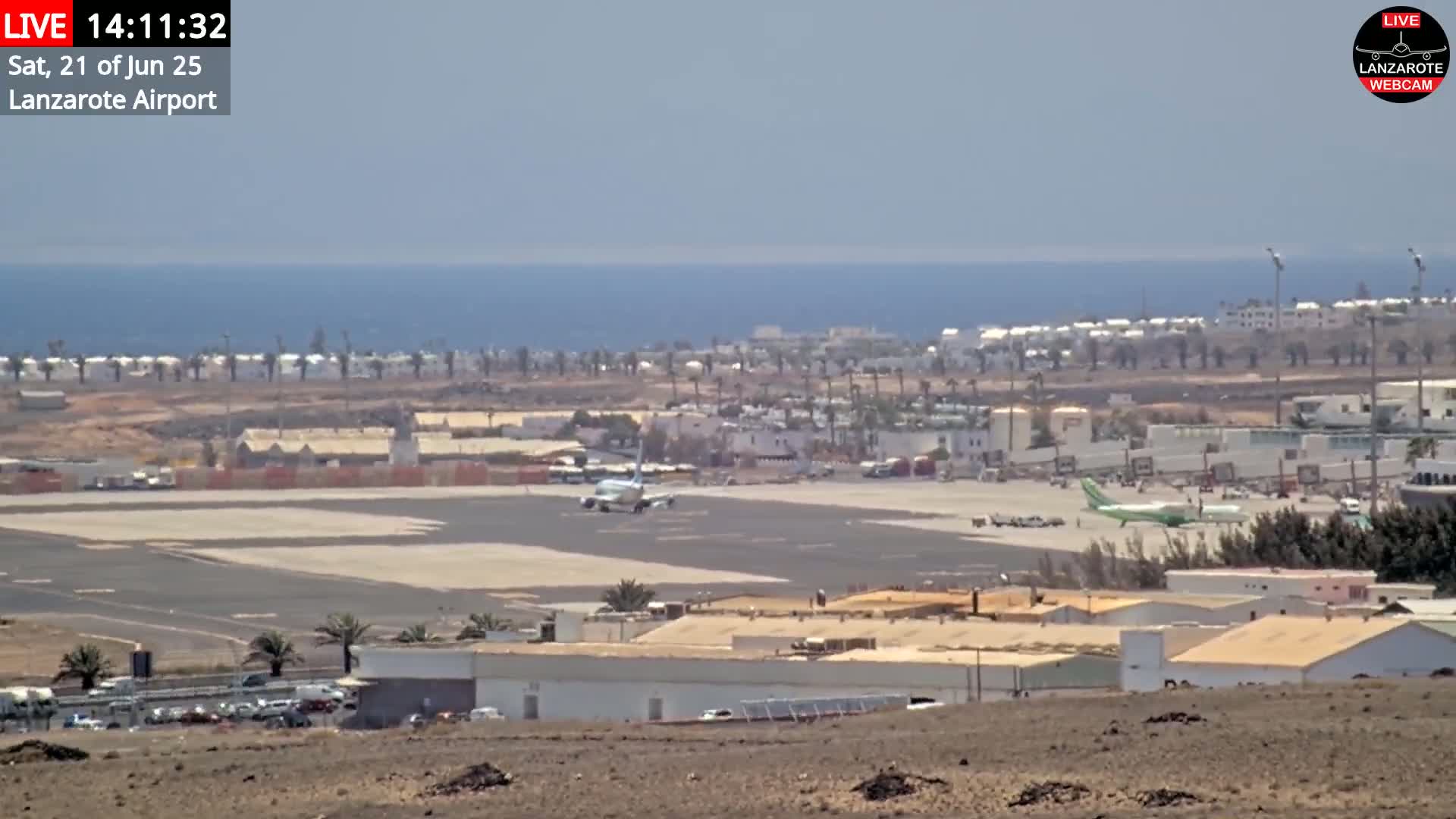 A sunny day at an airport shows a plane on the runway with buildings and a body of water in the background.