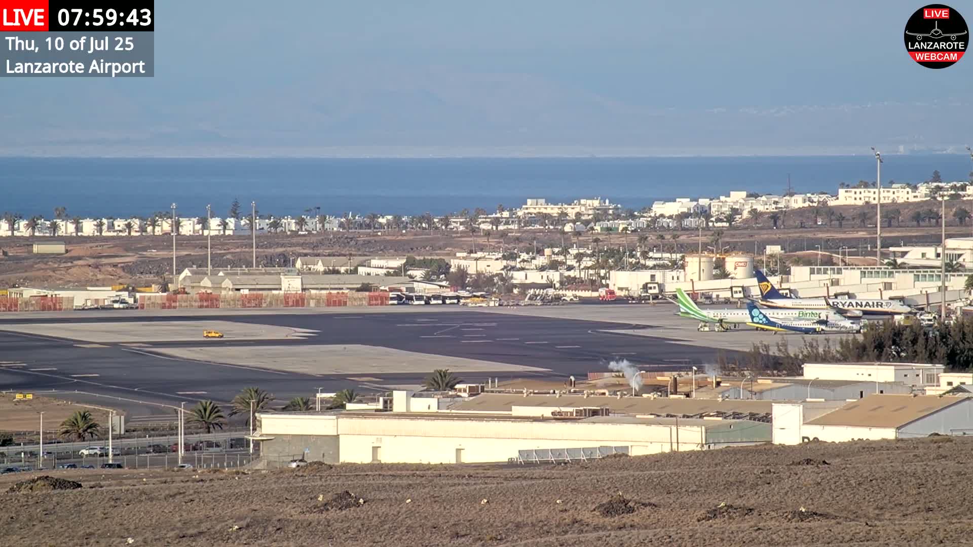 A clear day at an airport shows several planes parked on the tarmac with a body of water and buildings visible in the background.