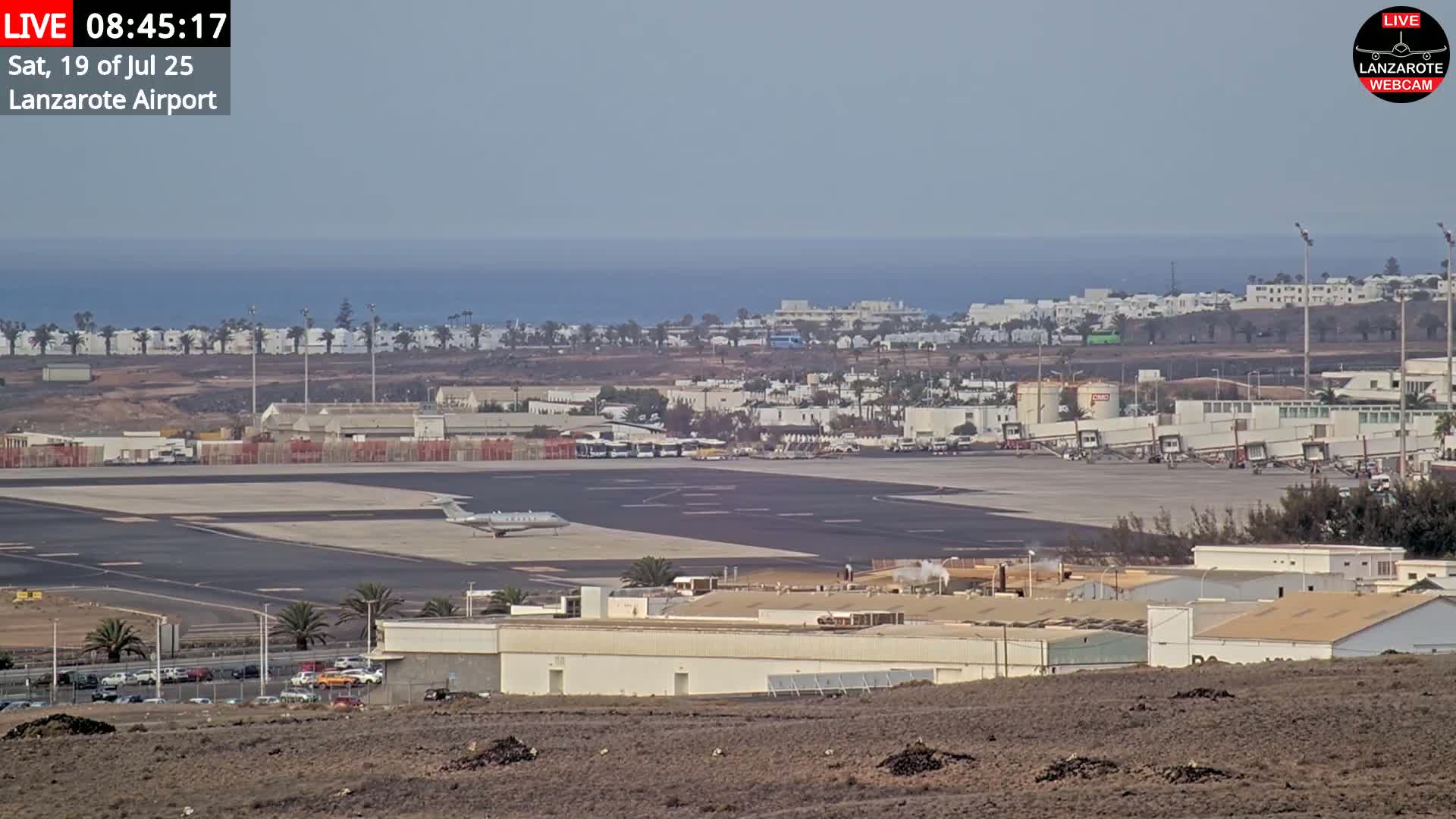 An airport tarmac with a private jet, surrounded by buildings and a distant ocean under hazy skies.