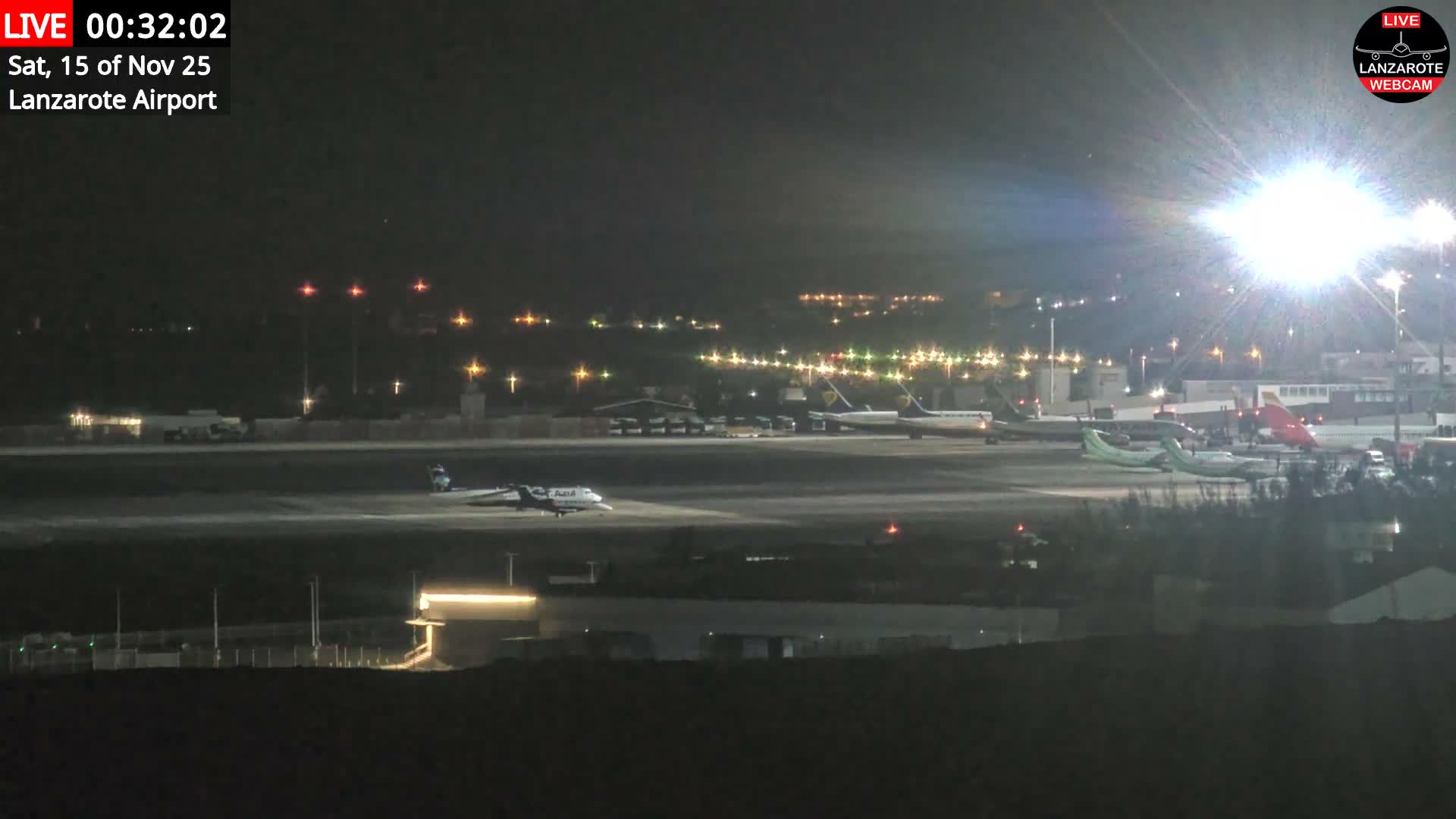 A clear night view of an airport tarmac shows multiple airplanes parked and one taxiing, illuminated by bright airport lights against a dark sky.