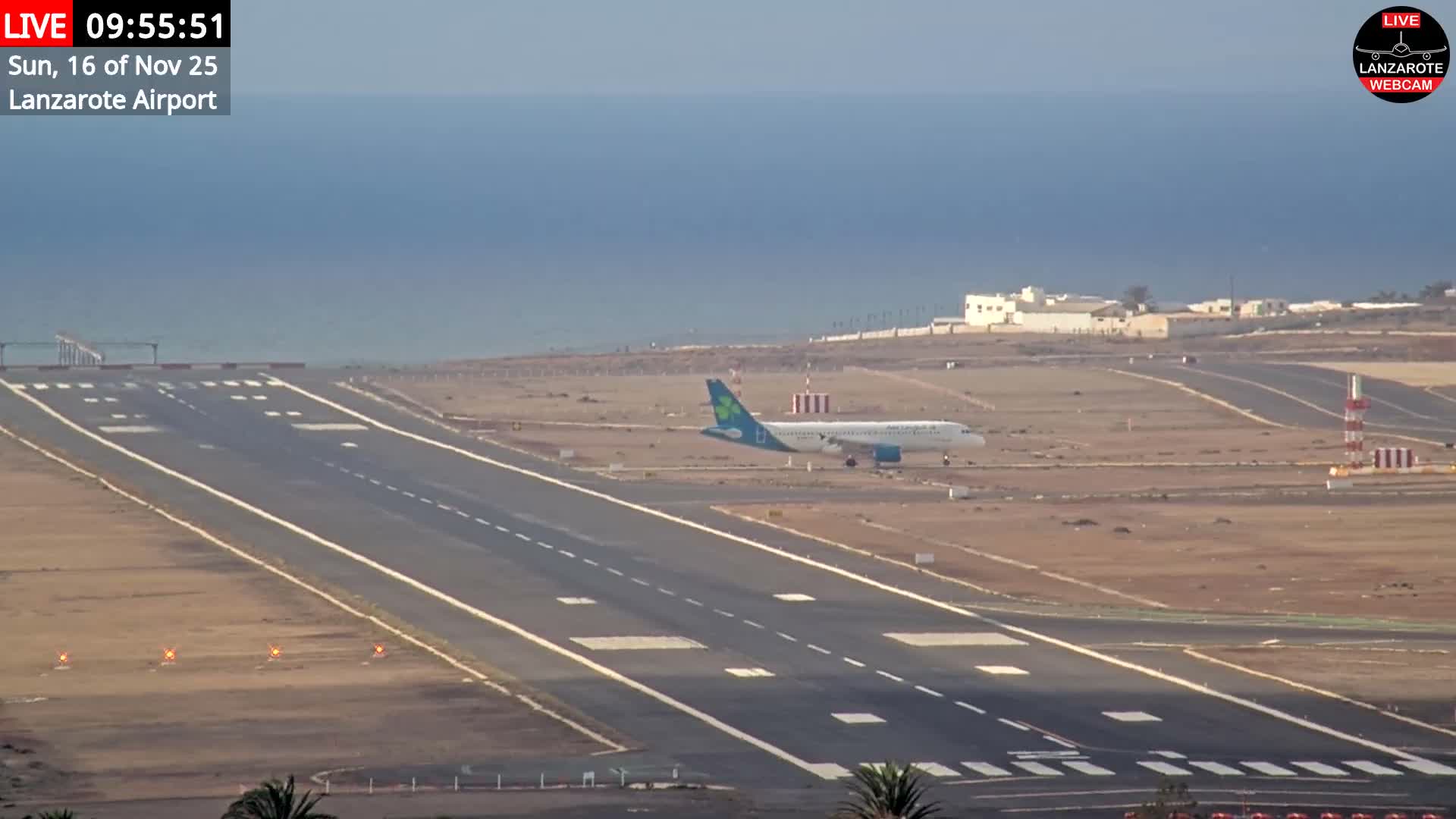 An Aer Lingus airplane is seen on a sunlit runway at an arid coastal airport, with the calm ocean visible in the background under clear skies.