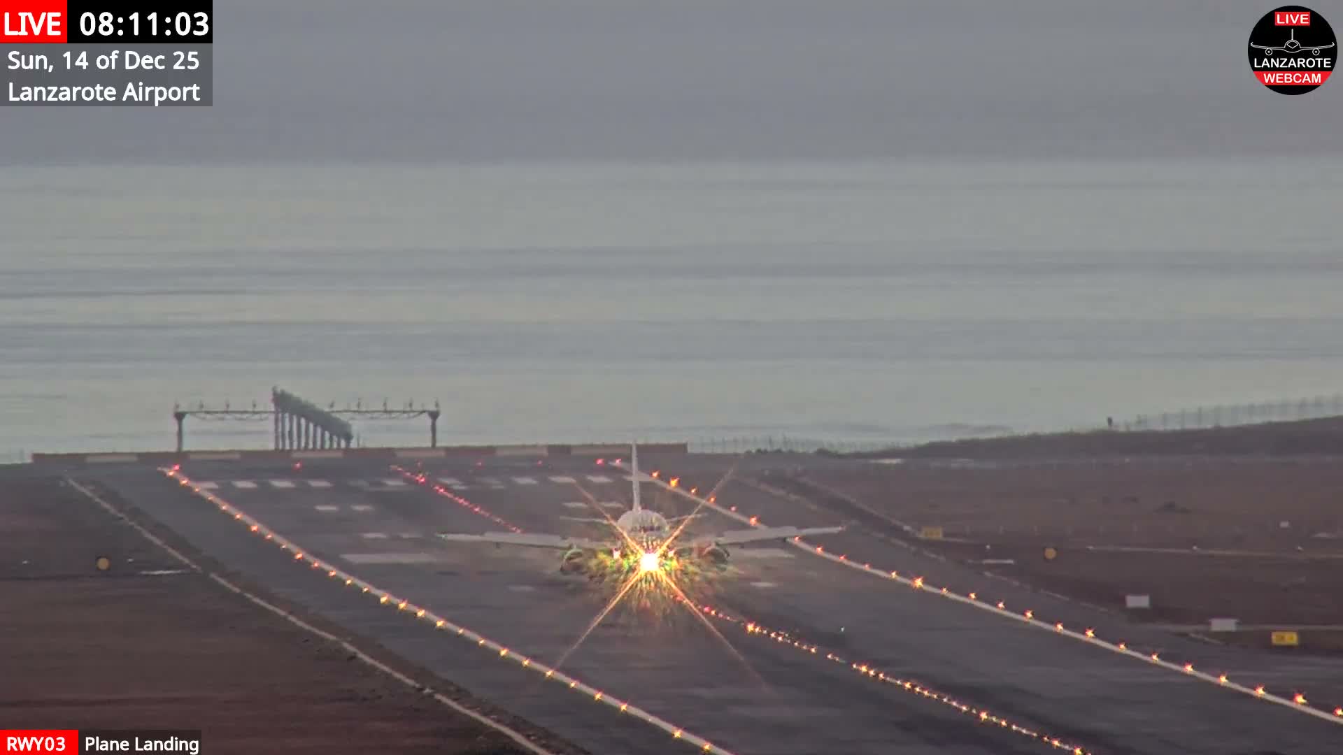 A commercial airplane with bright, starburst landing lights is centered on an airport runway next to the ocean under a gloomy, overcast sky.