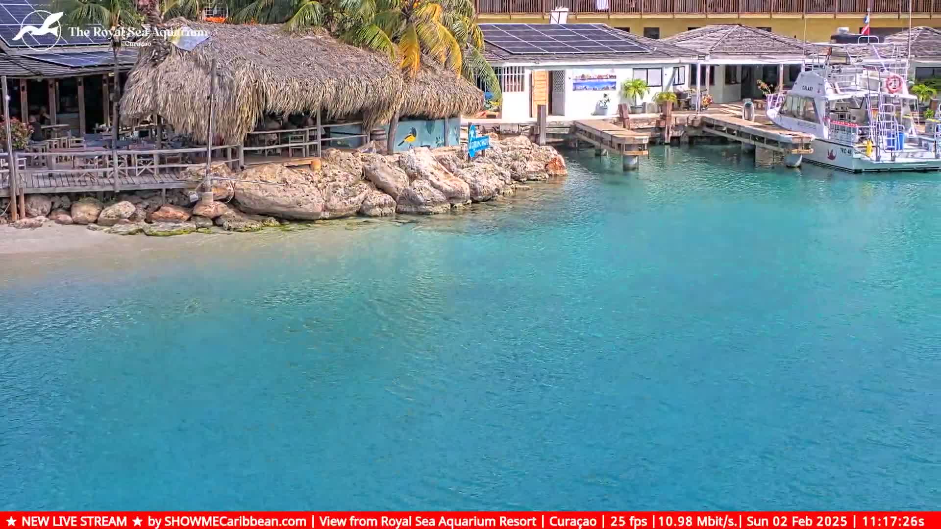 A sunny day shows a thatched-roof beachfront restaurant and a docked boat in clear turquoise water.