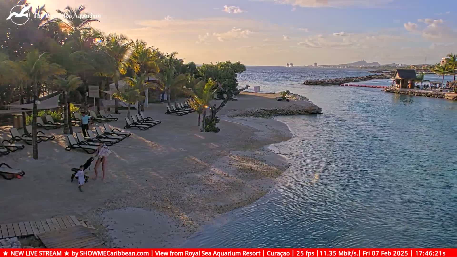 A sandy beach with lounge chairs and people is nestled next to a calm, clear body of water under a partly cloudy sky.