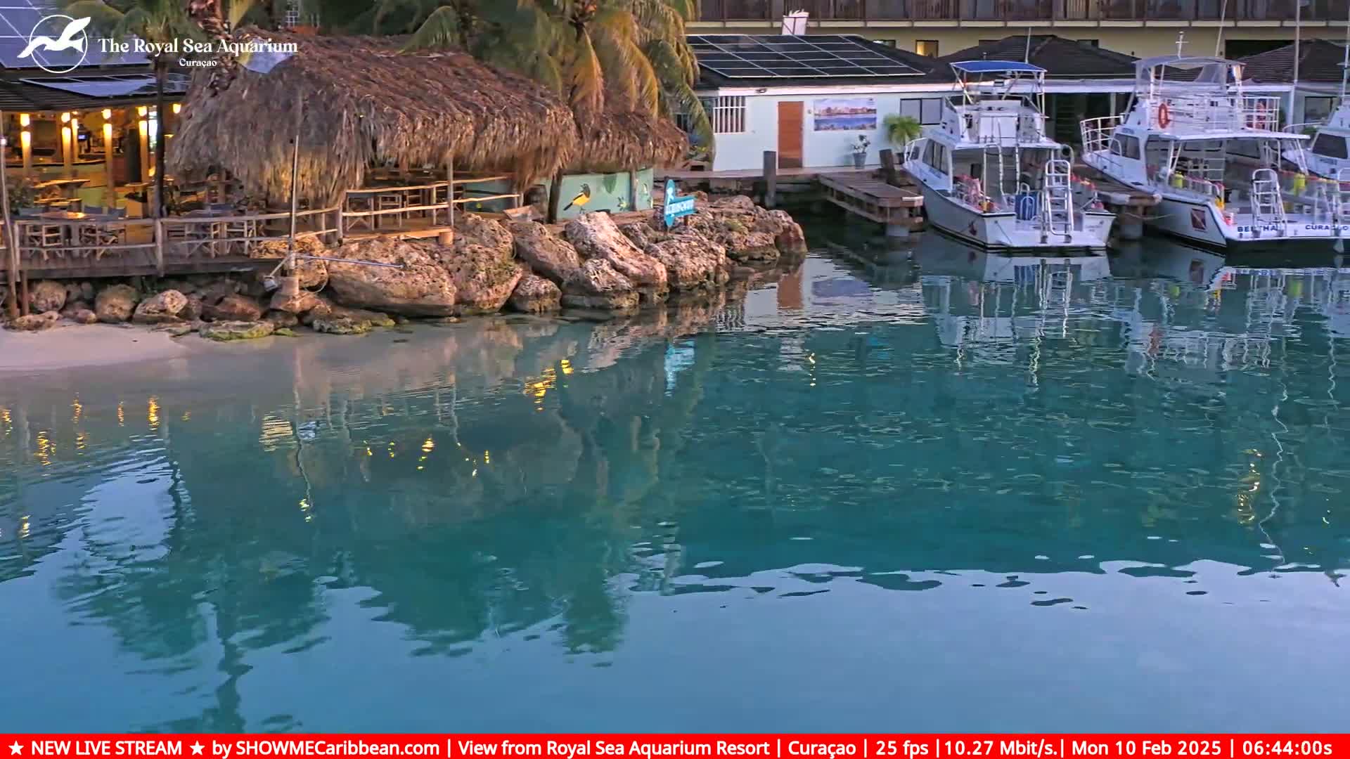 A tranquil waterfront scene at dusk shows a thatched-roof bar, a rocky shoreline, and several moored boats reflected in calm, teal water.