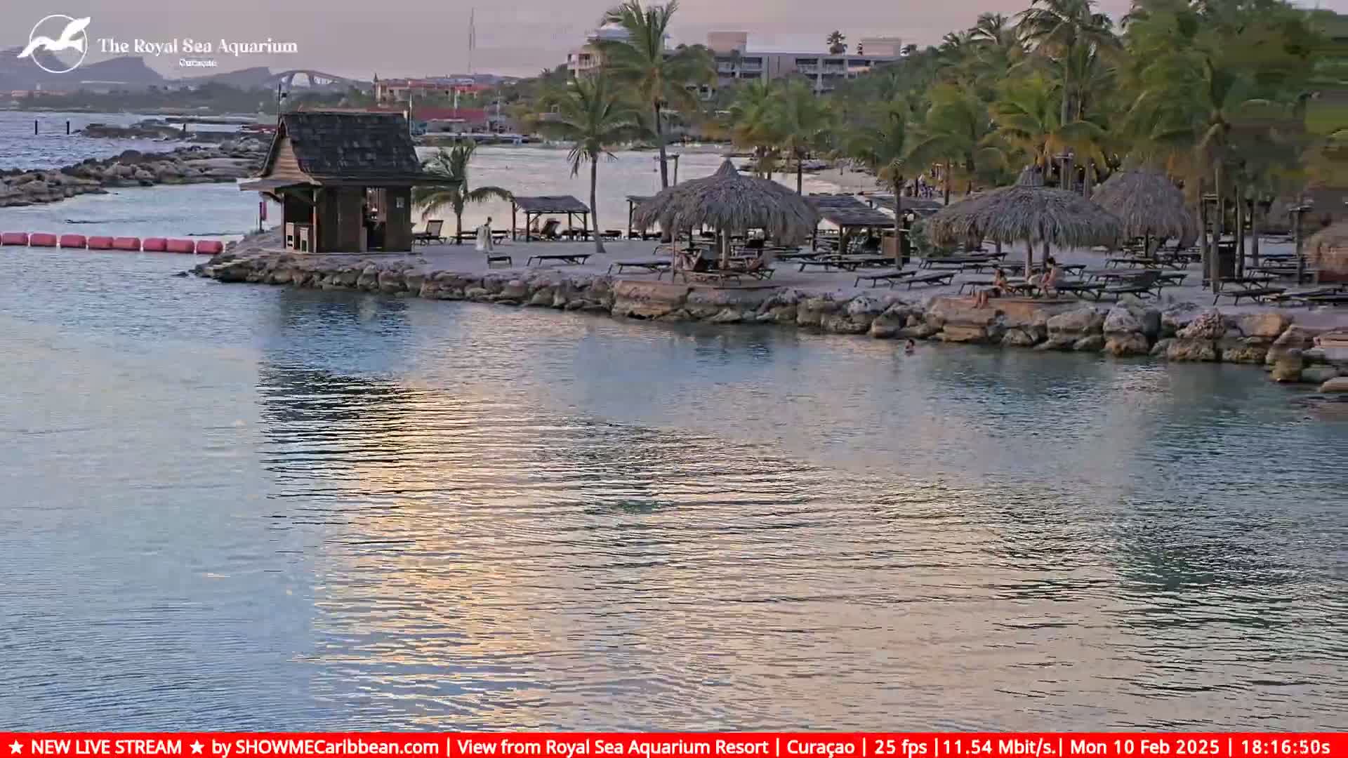 A calm body of water reflects the sunset, with a sandy beach featuring thatch-roofed umbrellas and lounge chairs in the background, under a mostly clear sky.