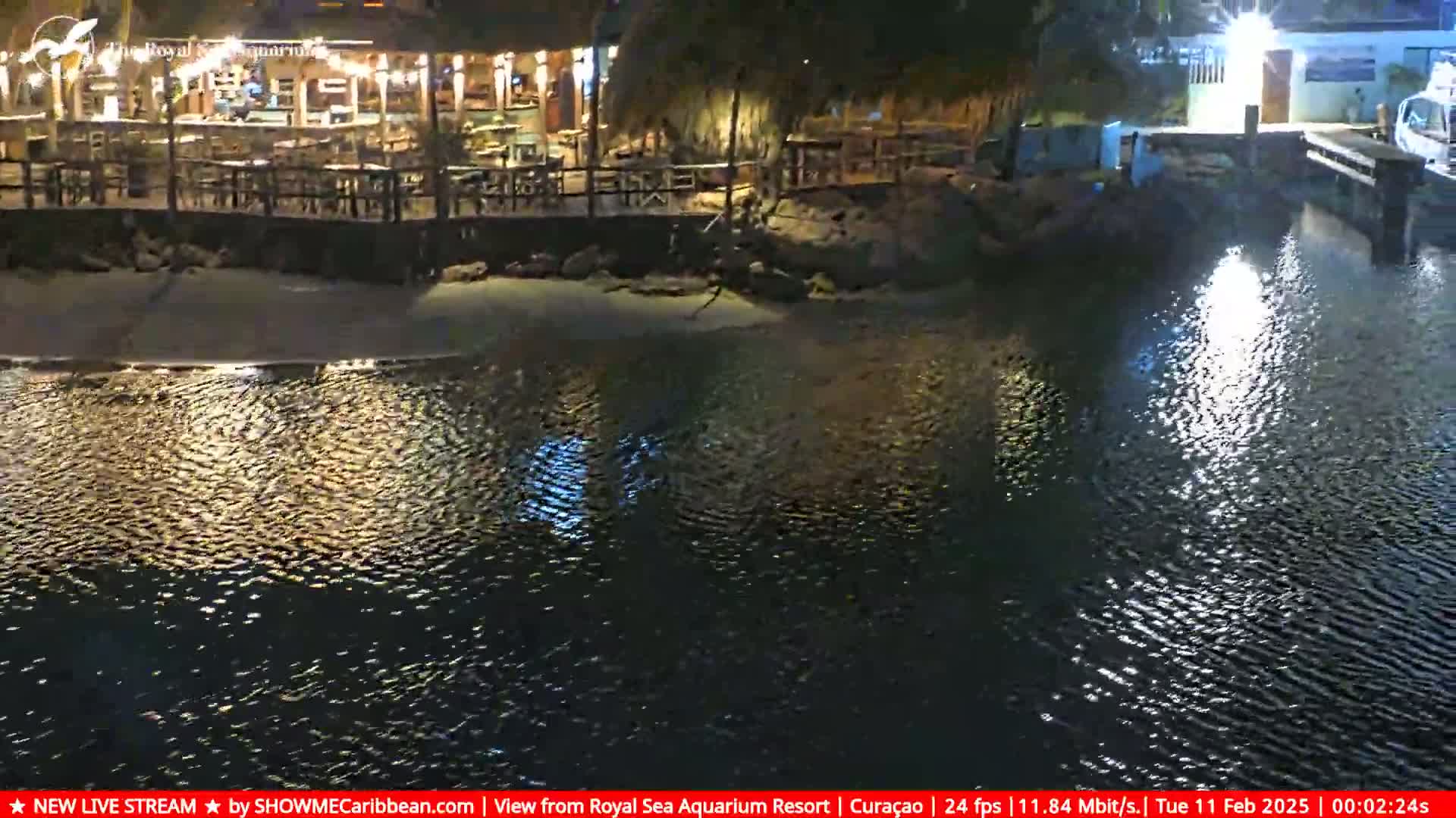 A nighttime view of a waterfront restaurant with outdoor seating illuminated by lights, next to calm, dark water reflecting the lights.