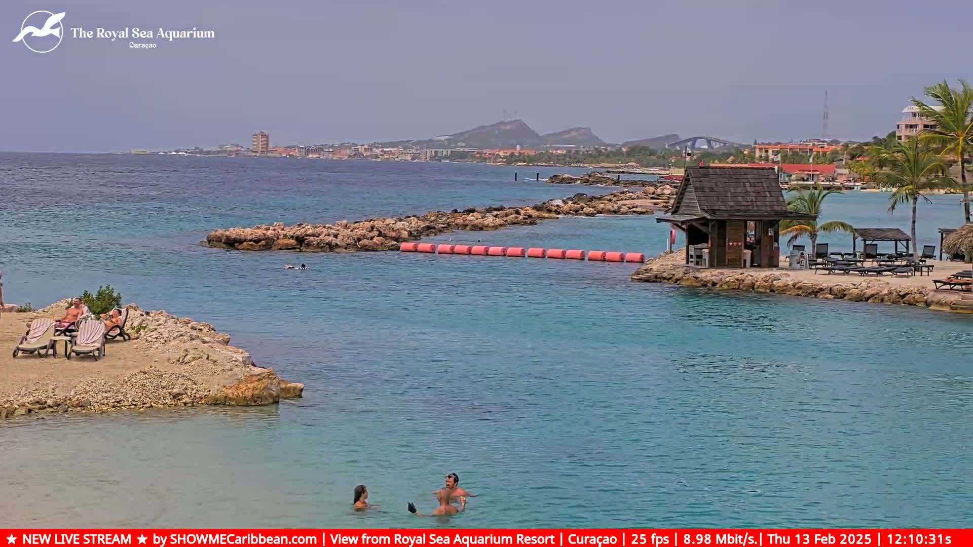 A calm, turquoise ocean scene features a rocky shoreline with lounge chairs and a small wooden hut, a floating barrier, and distant hills and buildings under a mostly cloudy sky.