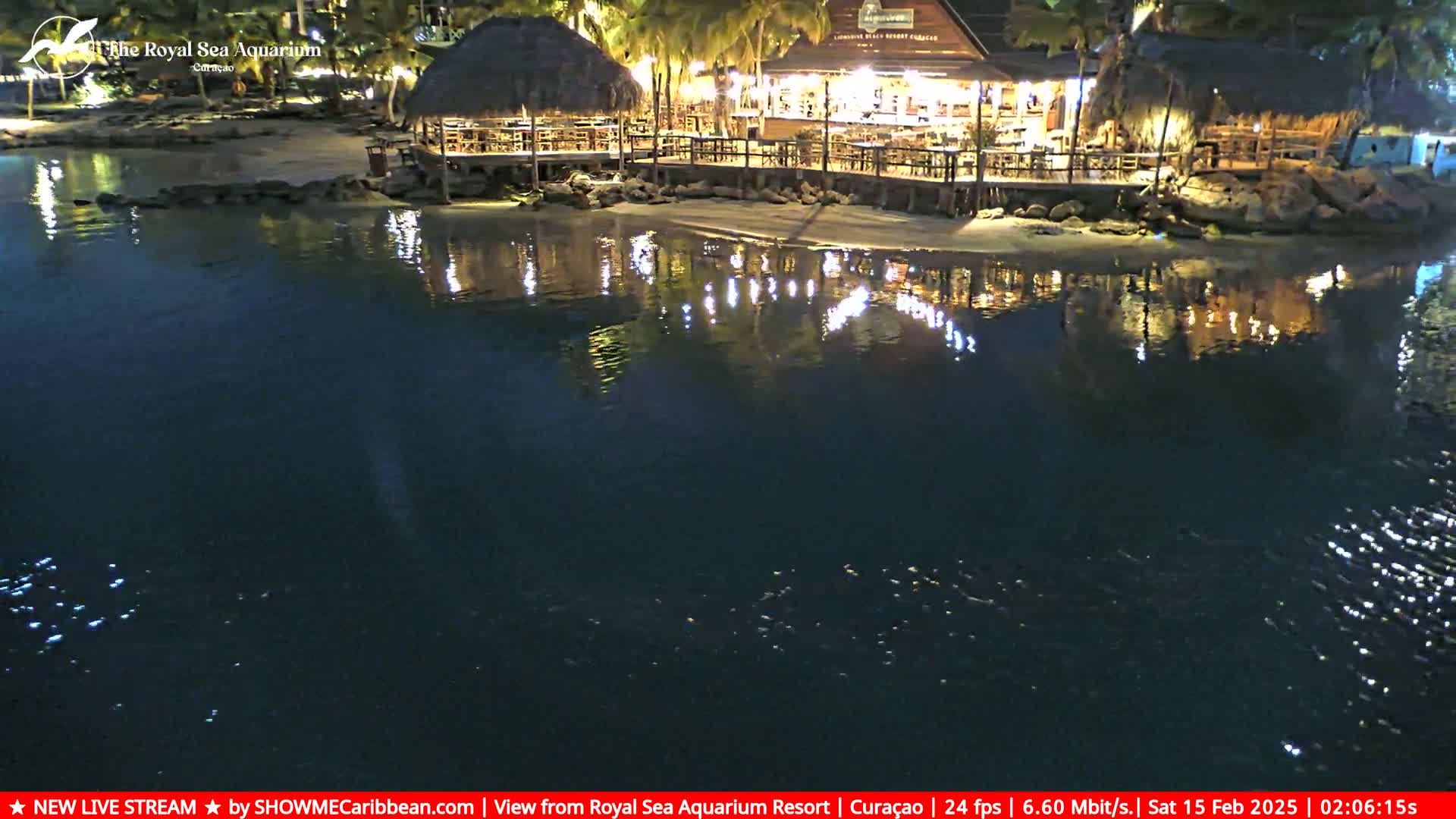 A waterfront restaurant with thatched roofs is brightly lit at night, its reflection shimmering on the dark water.