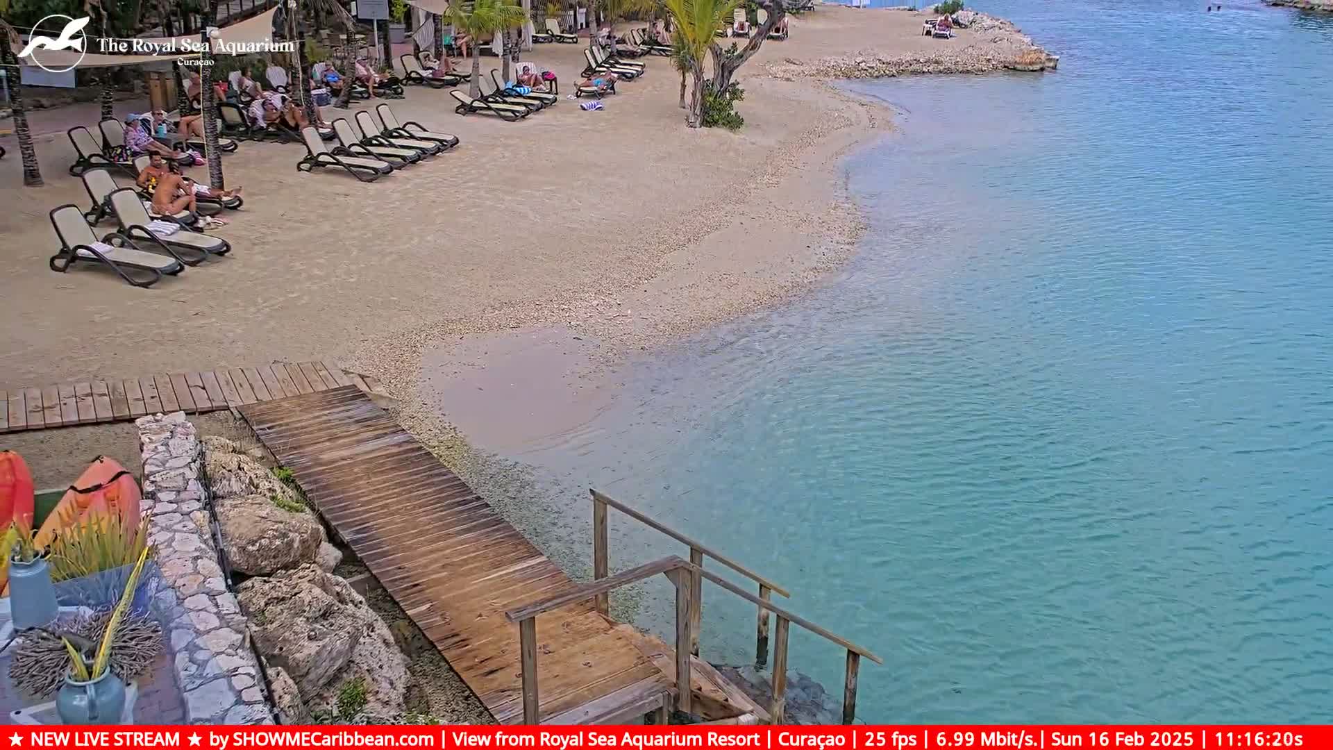 A sandy beach with lounge chairs and people relaxing, next to a calm, clear turquoise body of water with a wooden dock leading into it.