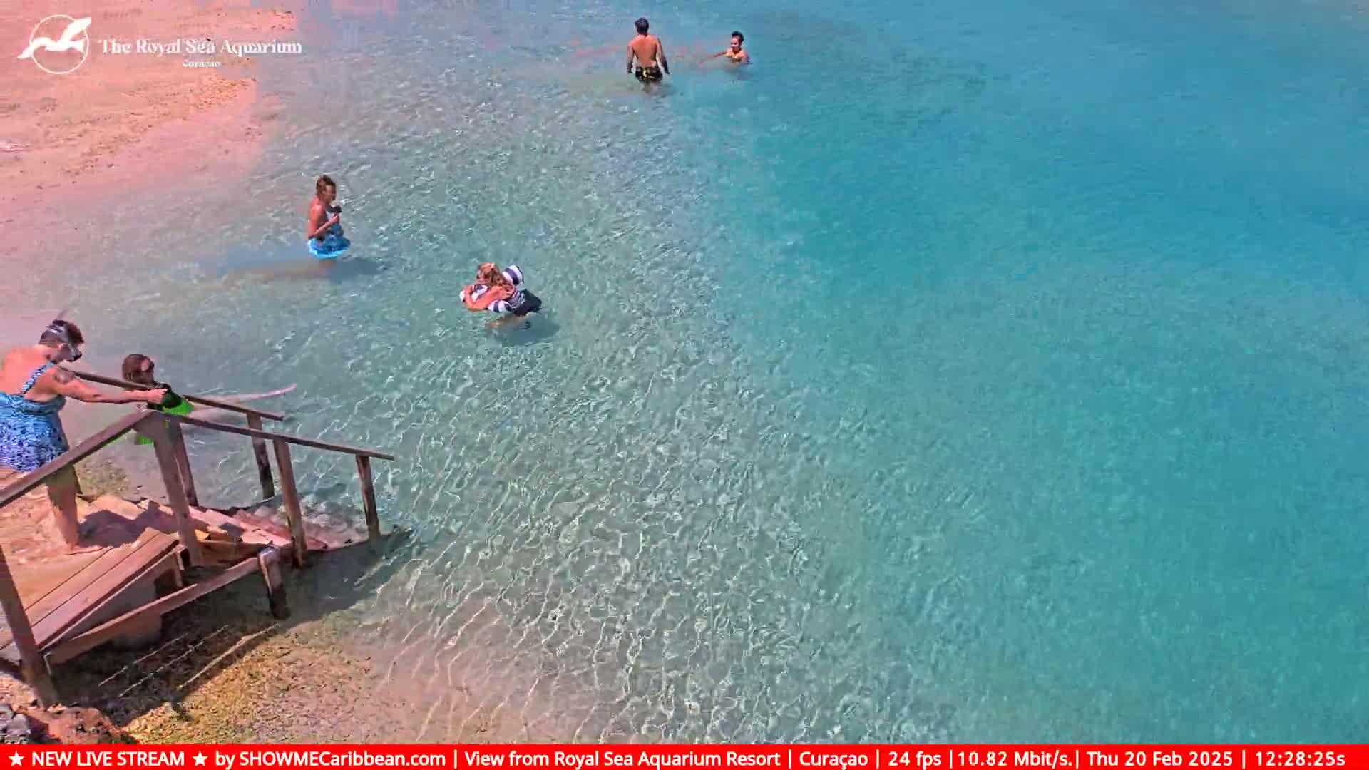Several people are wading and swimming in clear, shallow turquoise water near a wooden staircase leading to a sandy beach on a sunny day.