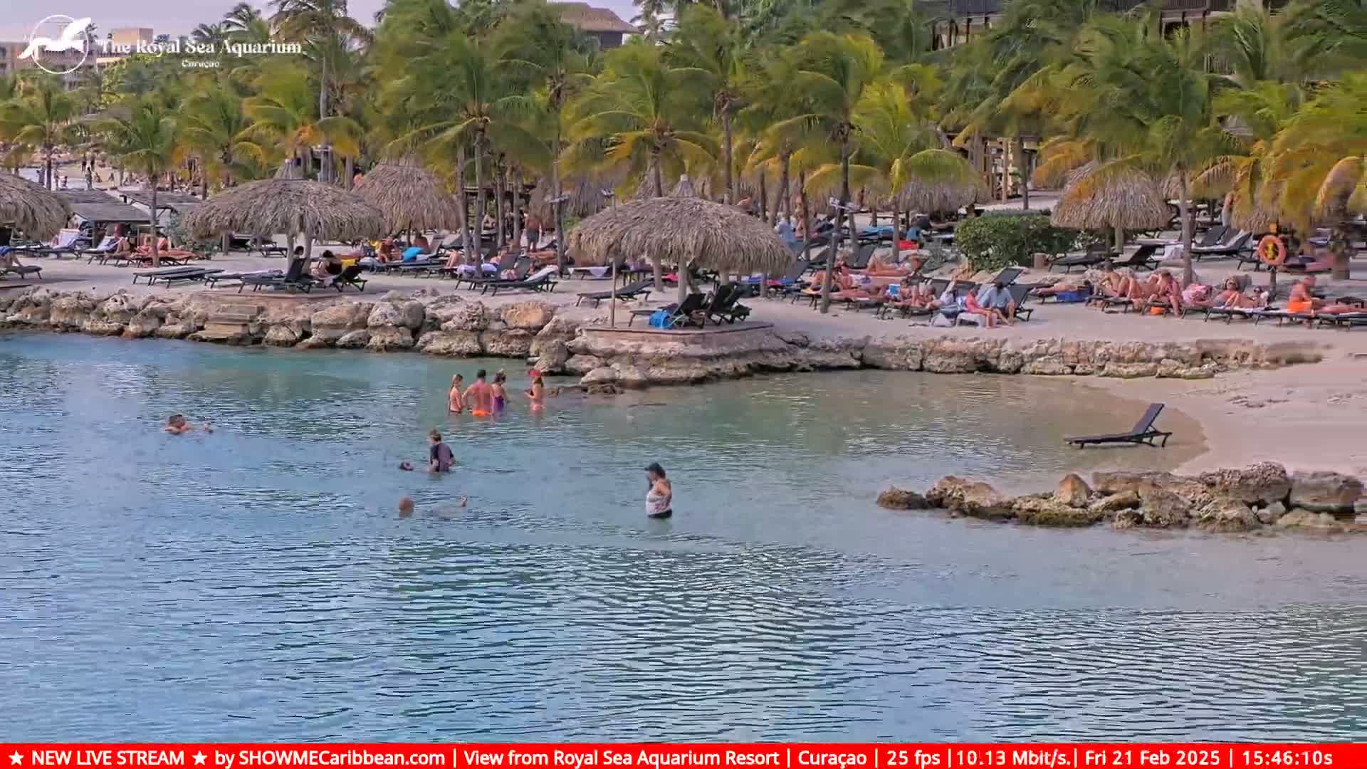 A calm, shallow body of water laps a sandy beach where people relax under thatched umbrellas and swim in the clear water, surrounded by palm trees.