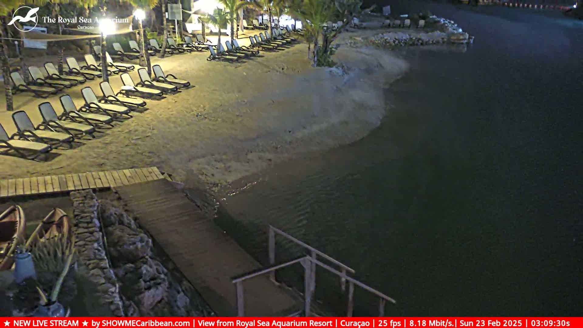 A sandy beach at night with empty lounge chairs next to a calm body of water and a wooden dock leading into the water.